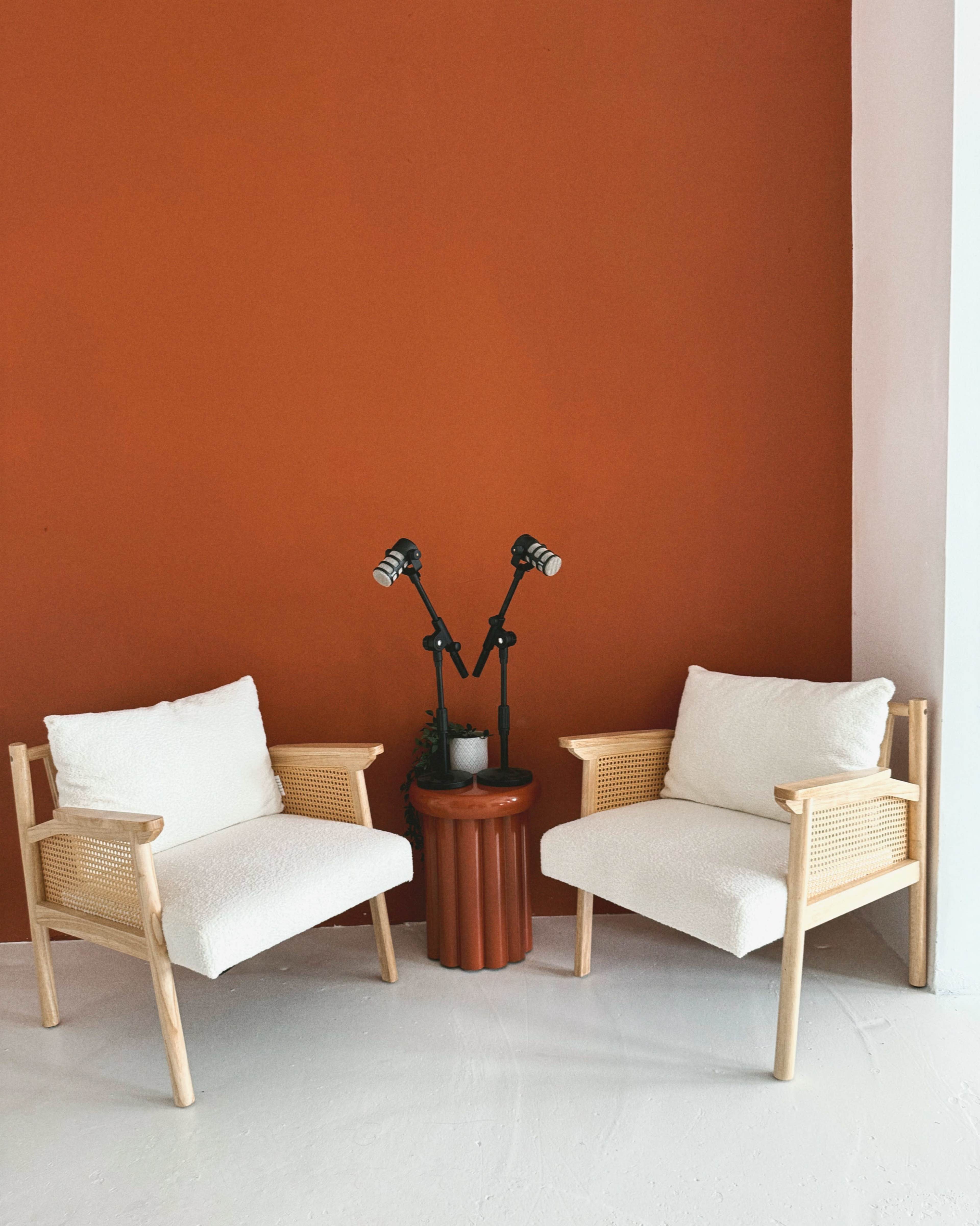 Two light-colored chairs with a wooden frame and plush white cushions are positioned opposite each other, flanking a small, cylindrical planter with a plant and two adjustable lamps, set against a solid orange wall.