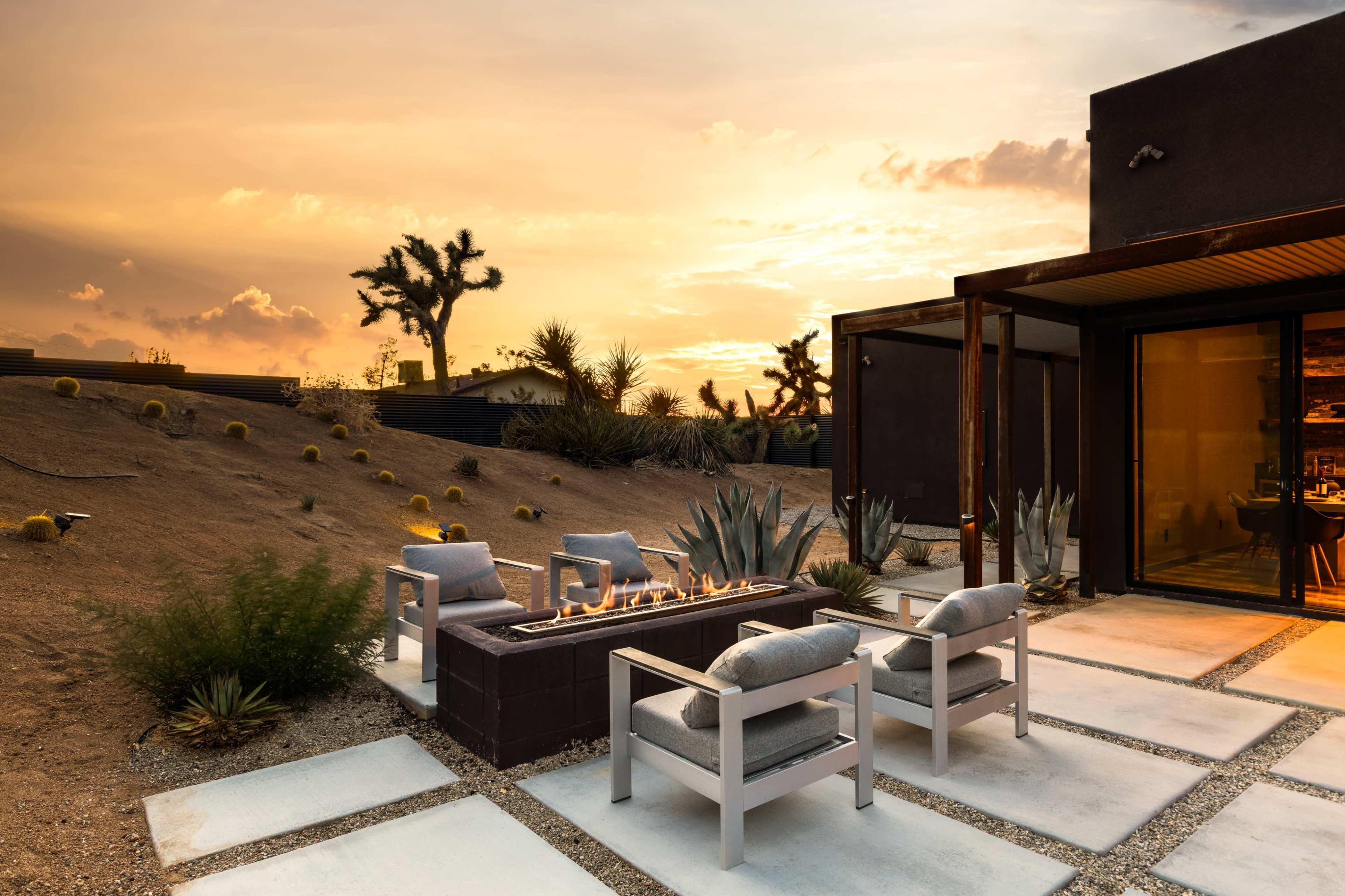 A modern outdoor seating area features four chairs arranged around a fire pit, set against a desert landscape at sunset.