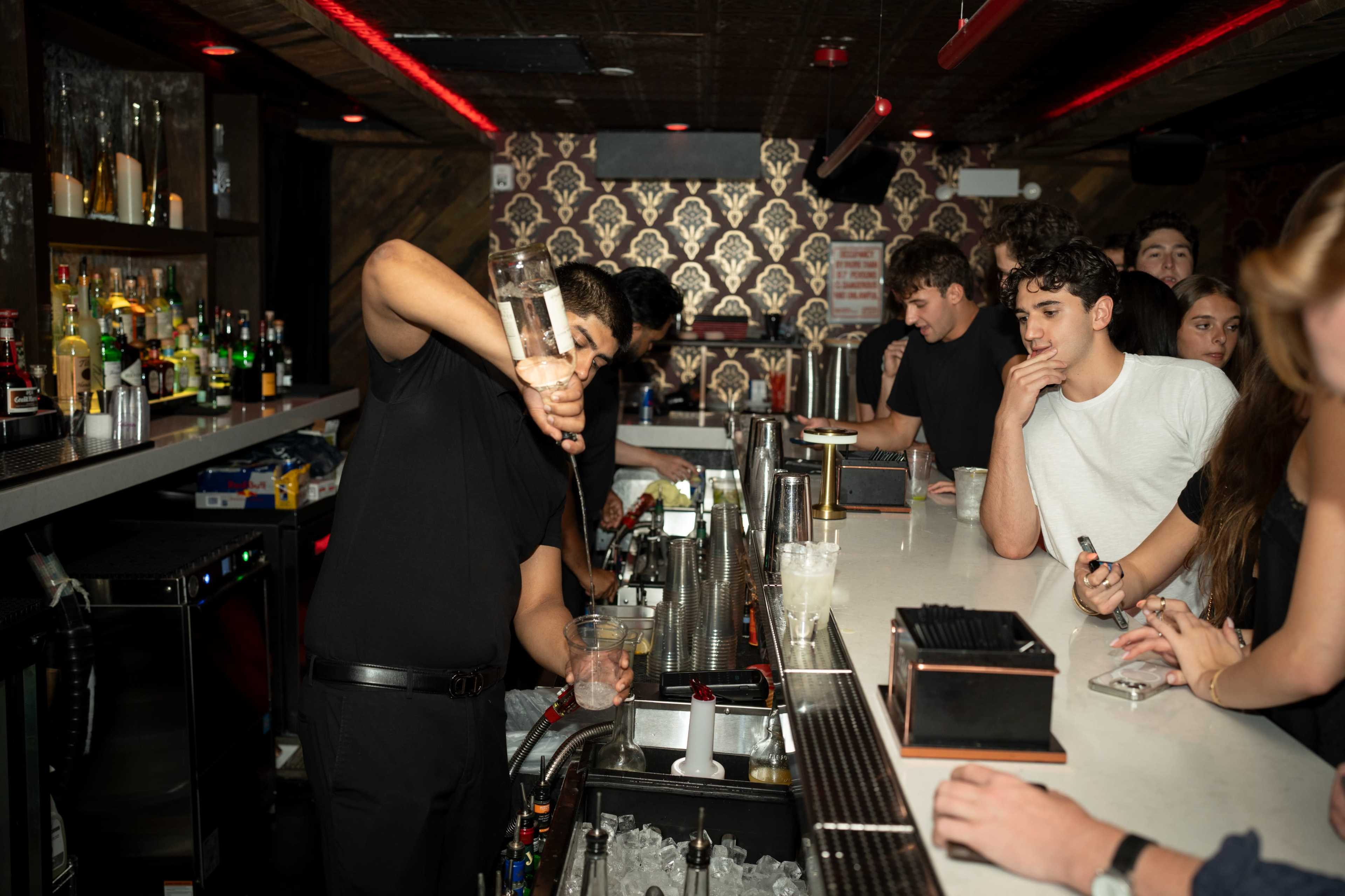A bartender prepares a cocktail at a bustling bar while customers watch with interest.