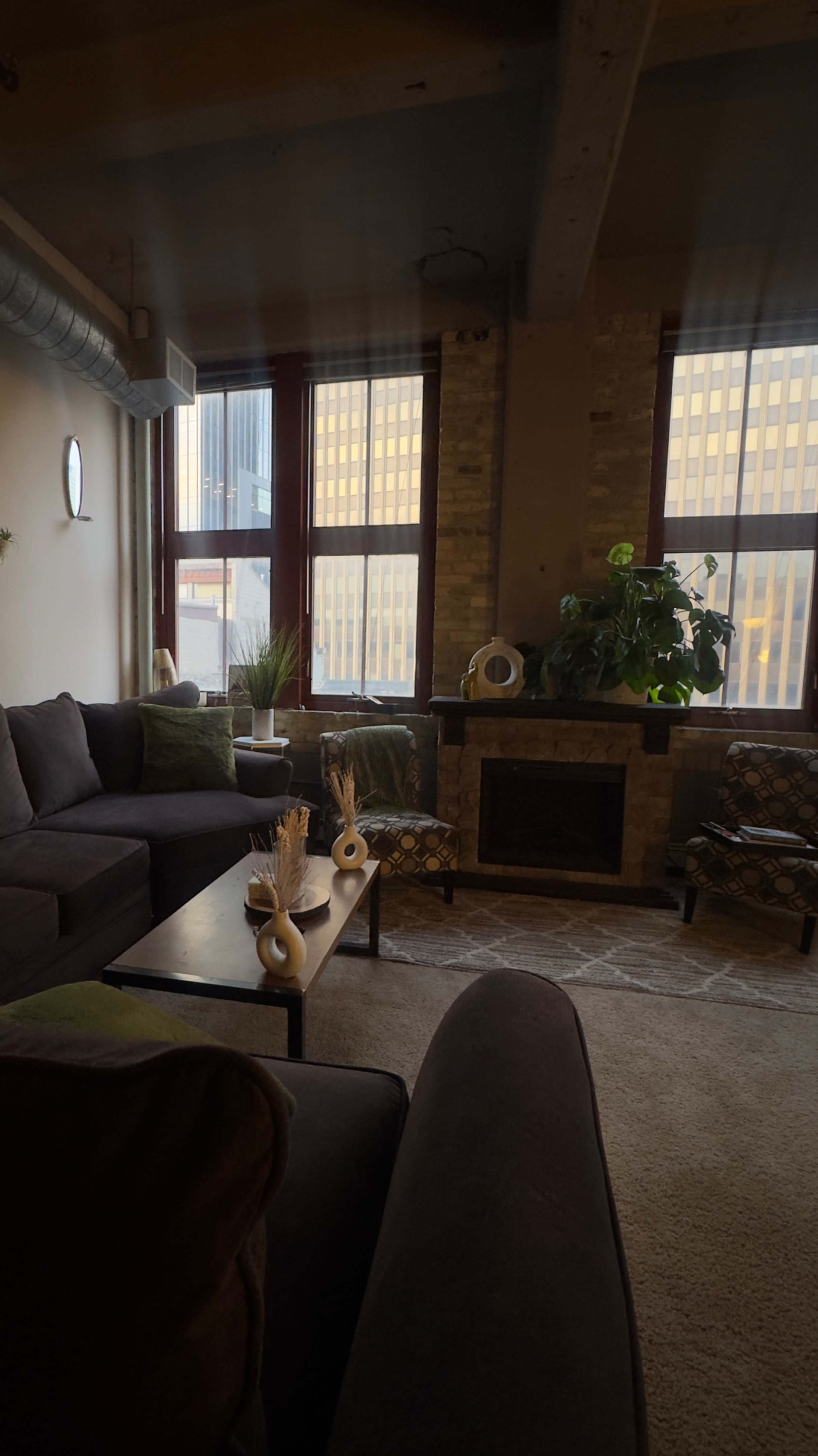 The image shows a cozy living room with large windows, a stone fireplace, and comfortable furniture arranged around a central coffee table.