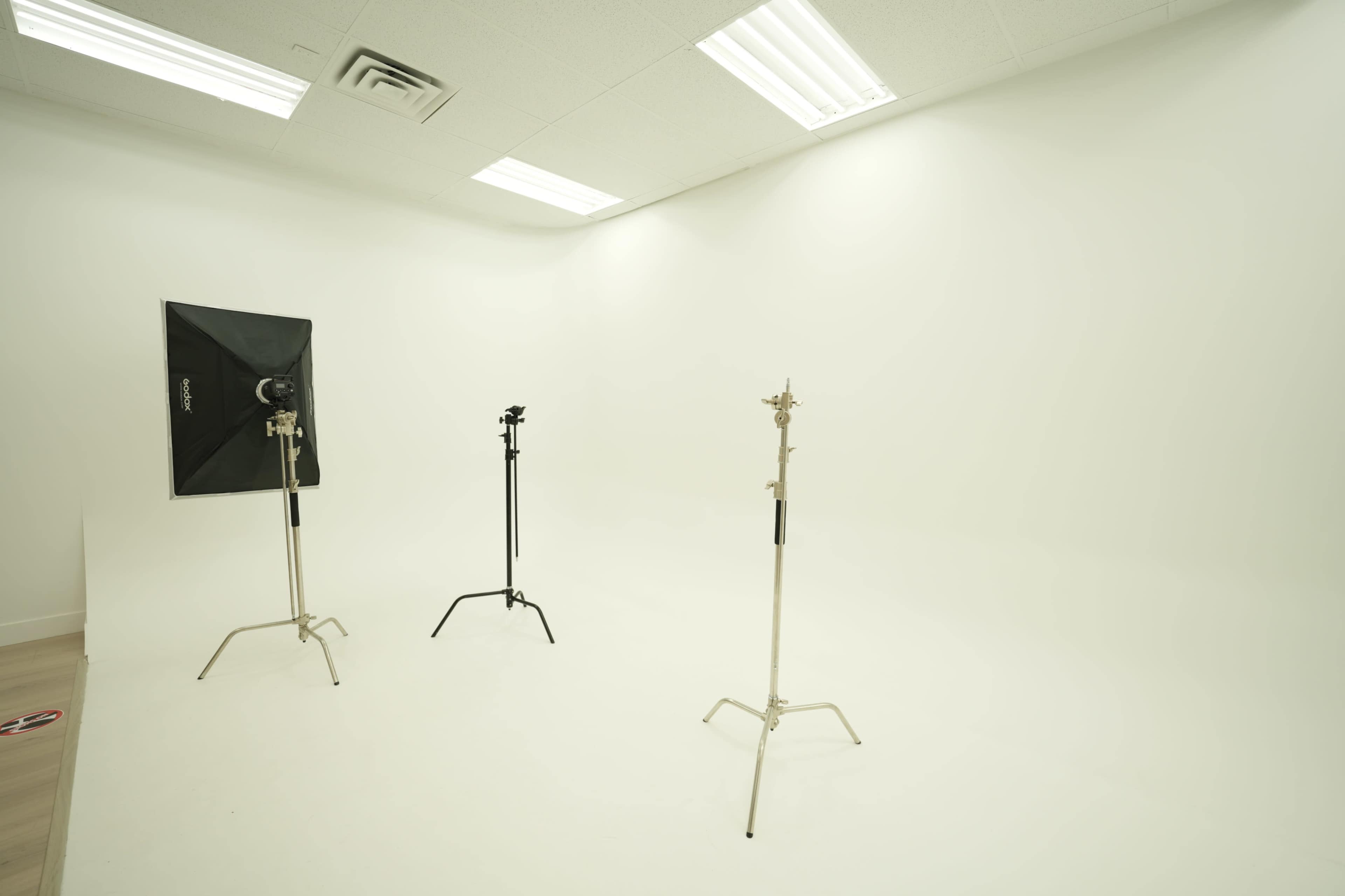 The image shows an empty photo studio with a plain white background and two light stands positioned in the center.