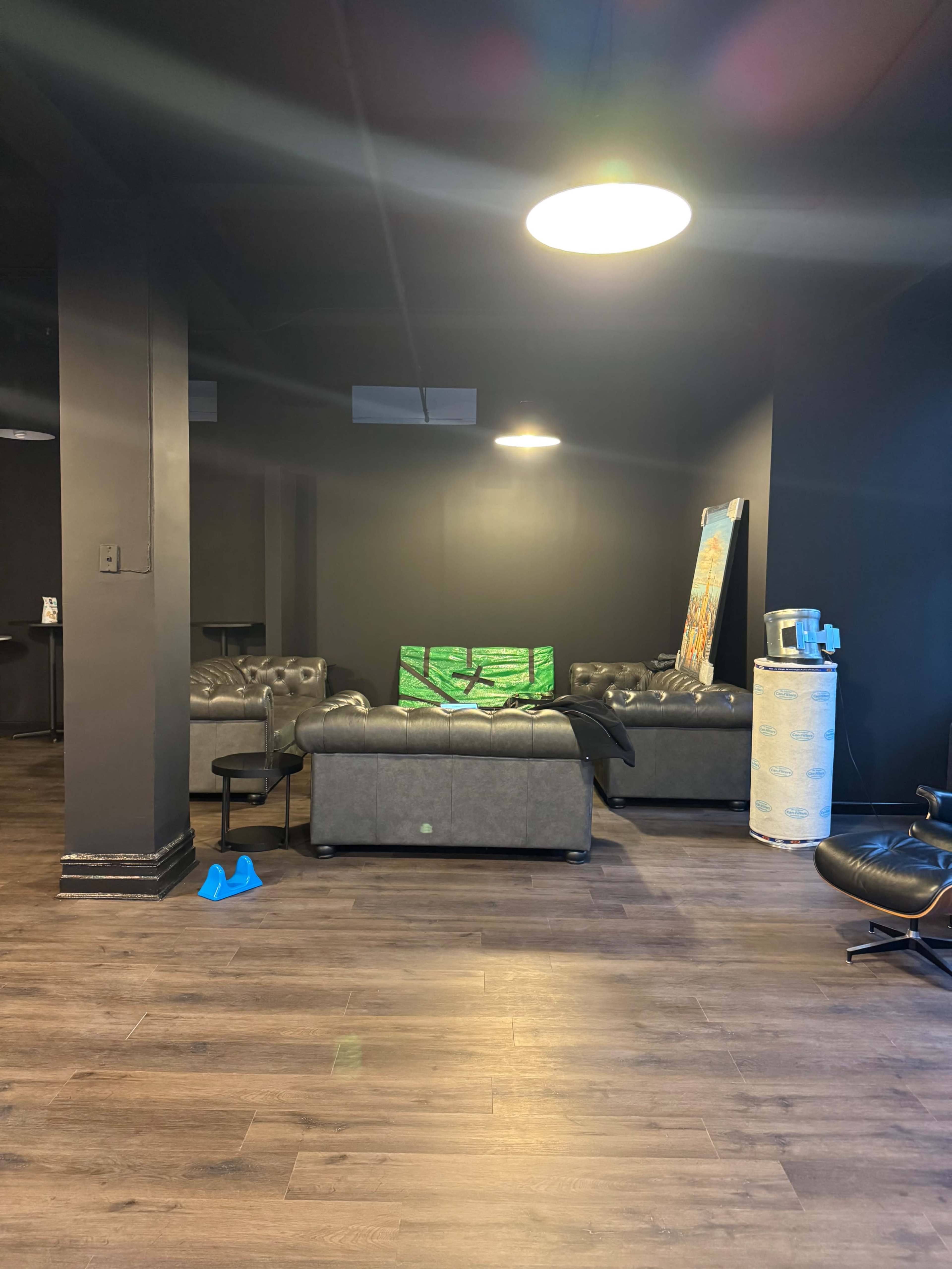 The image shows a dimly lit lounge area with a black sofa, a green canvas leaning against the wall, and a few scattered furniture pieces on a wooden floor.