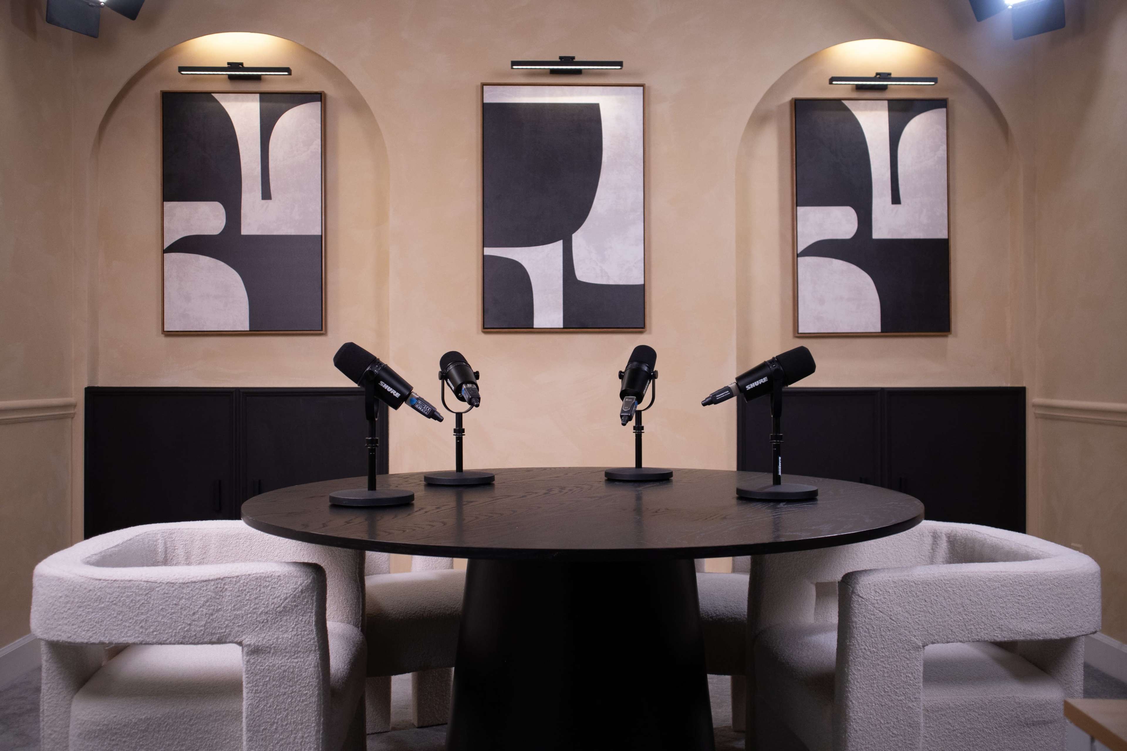 A modern studio setup with a round black table, three microphones positioned on it, and two white chairs, accompanied by abstract black and white artwork on the walls.
