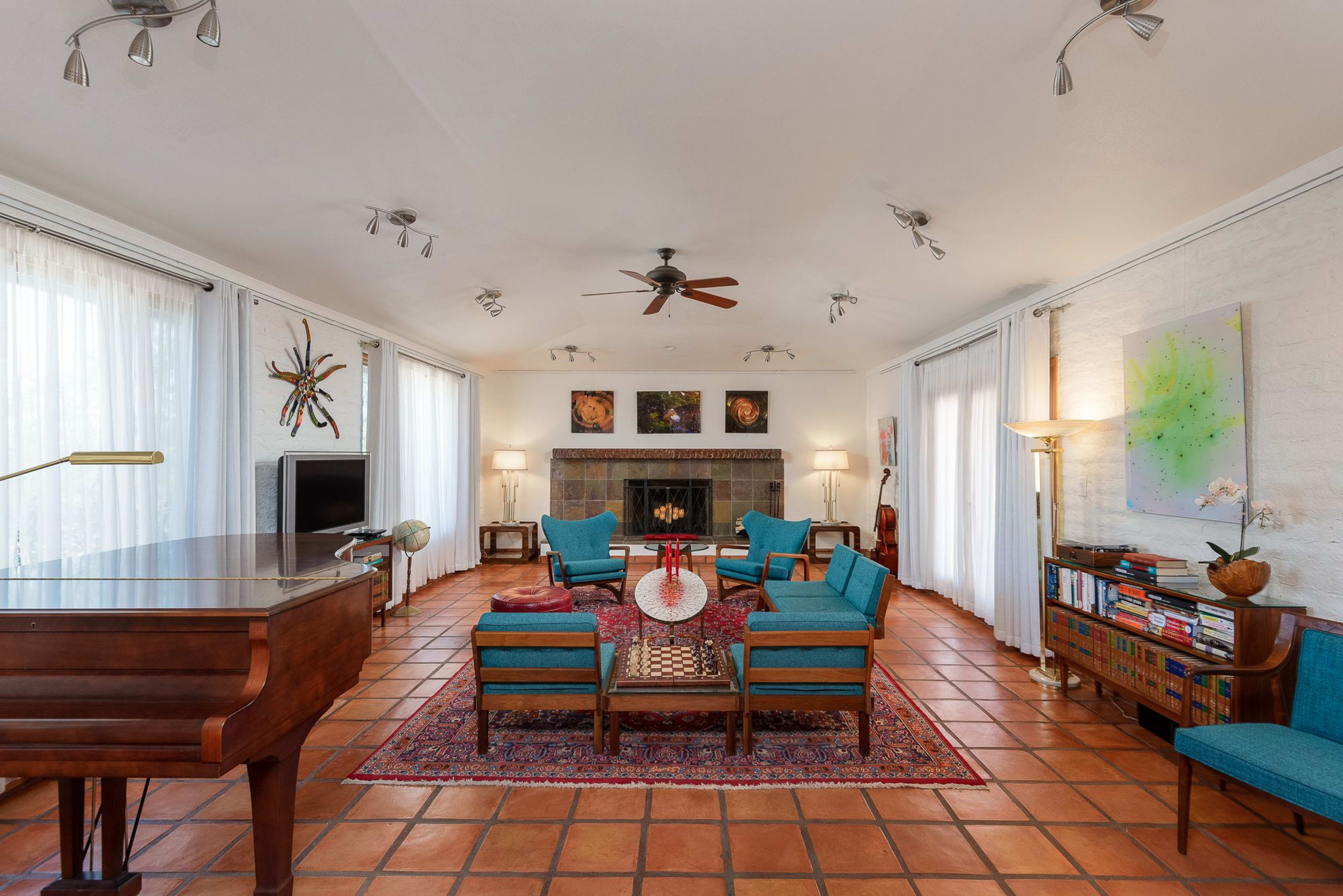 A spacious living room with a piano, a seating arrangement of teal chairs and a wooden table, and a fireplace surrounded by artwork and large windows letting in natural light.