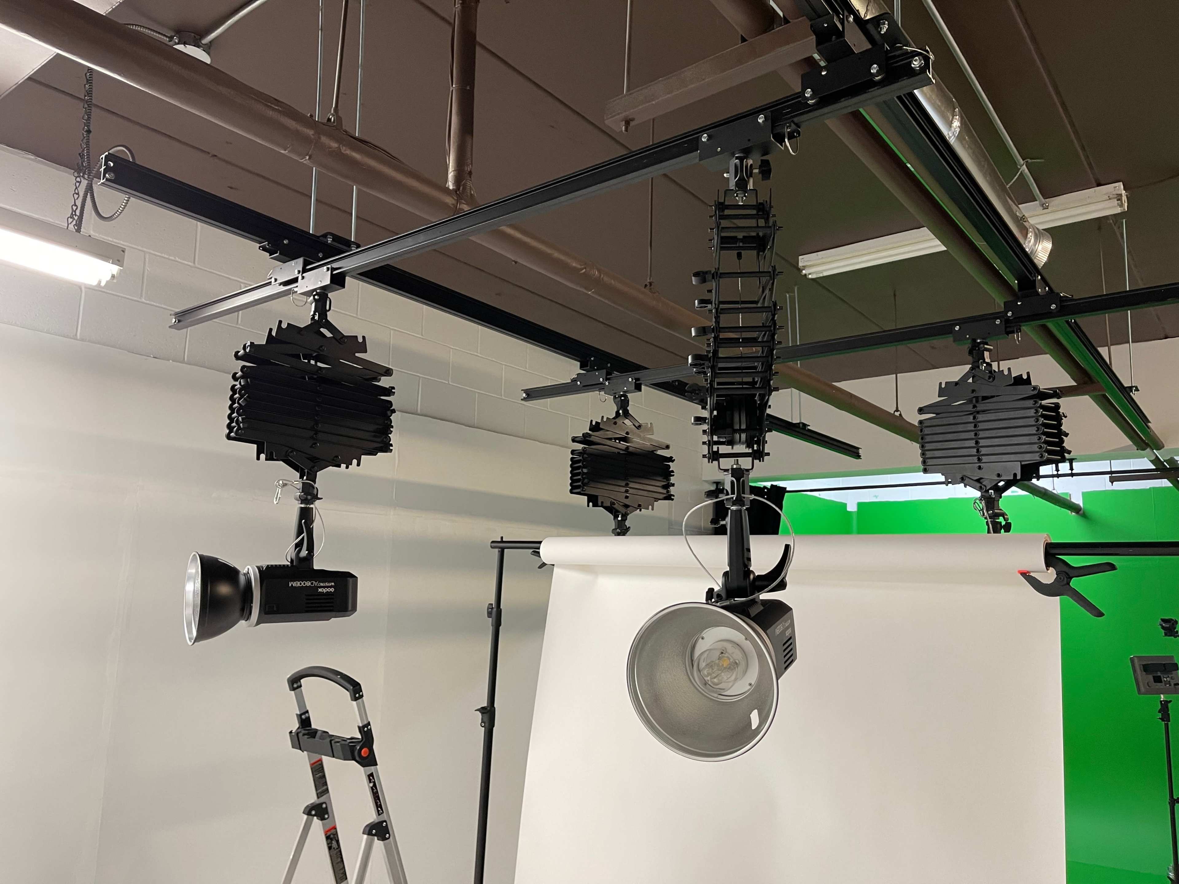 A lighting setup in a studio with multiple adjustable lights mounted on a track above a white backdrop and a green screen.