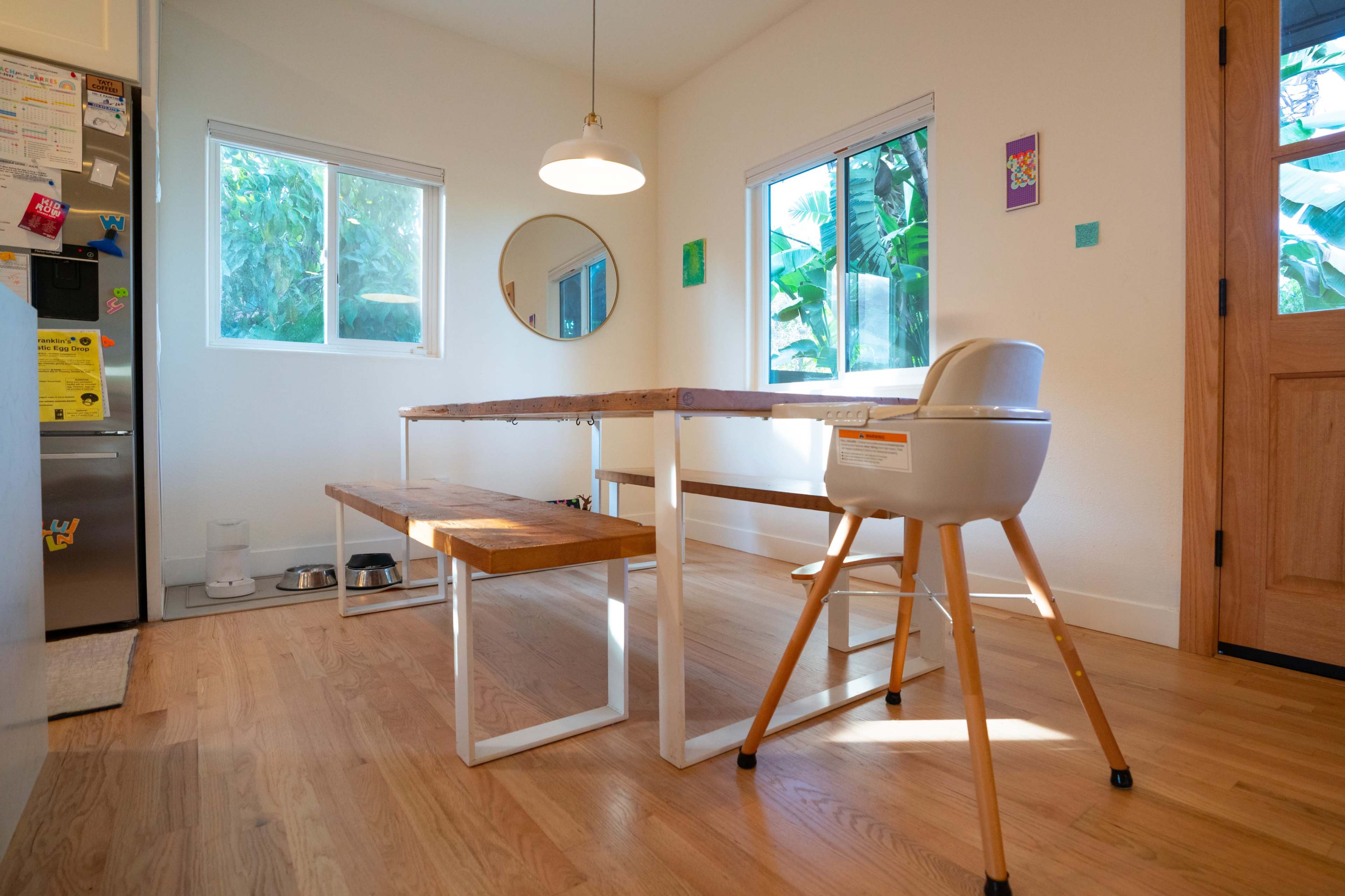 A modern dining area features a wooden table with benches, a high chair, and large windows allowing natural light to illuminate the space.