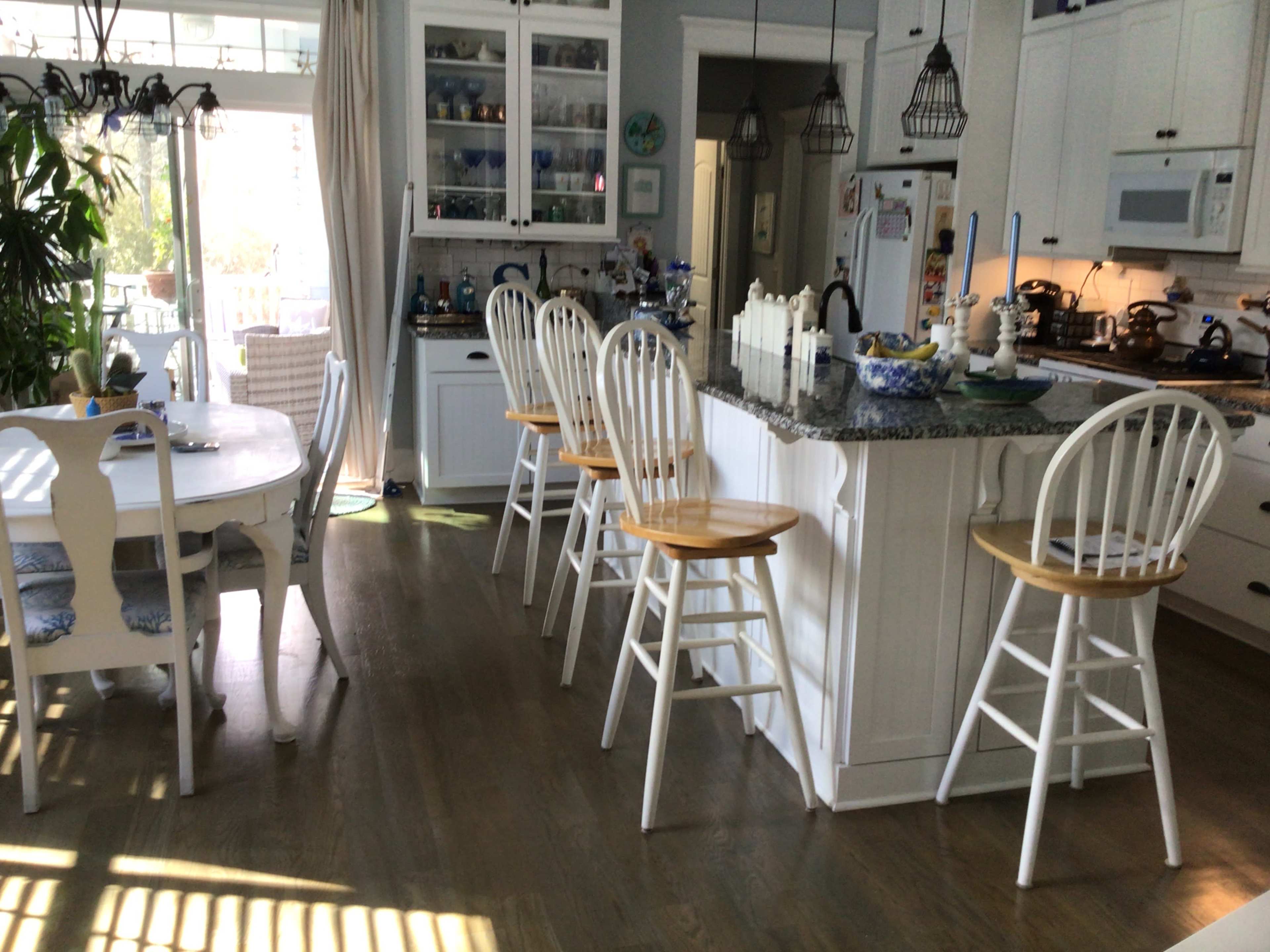 A bright kitchen with a granite countertop, white bar stools, and a dining table adjacent to it, featuring natural light streaming through large windows.