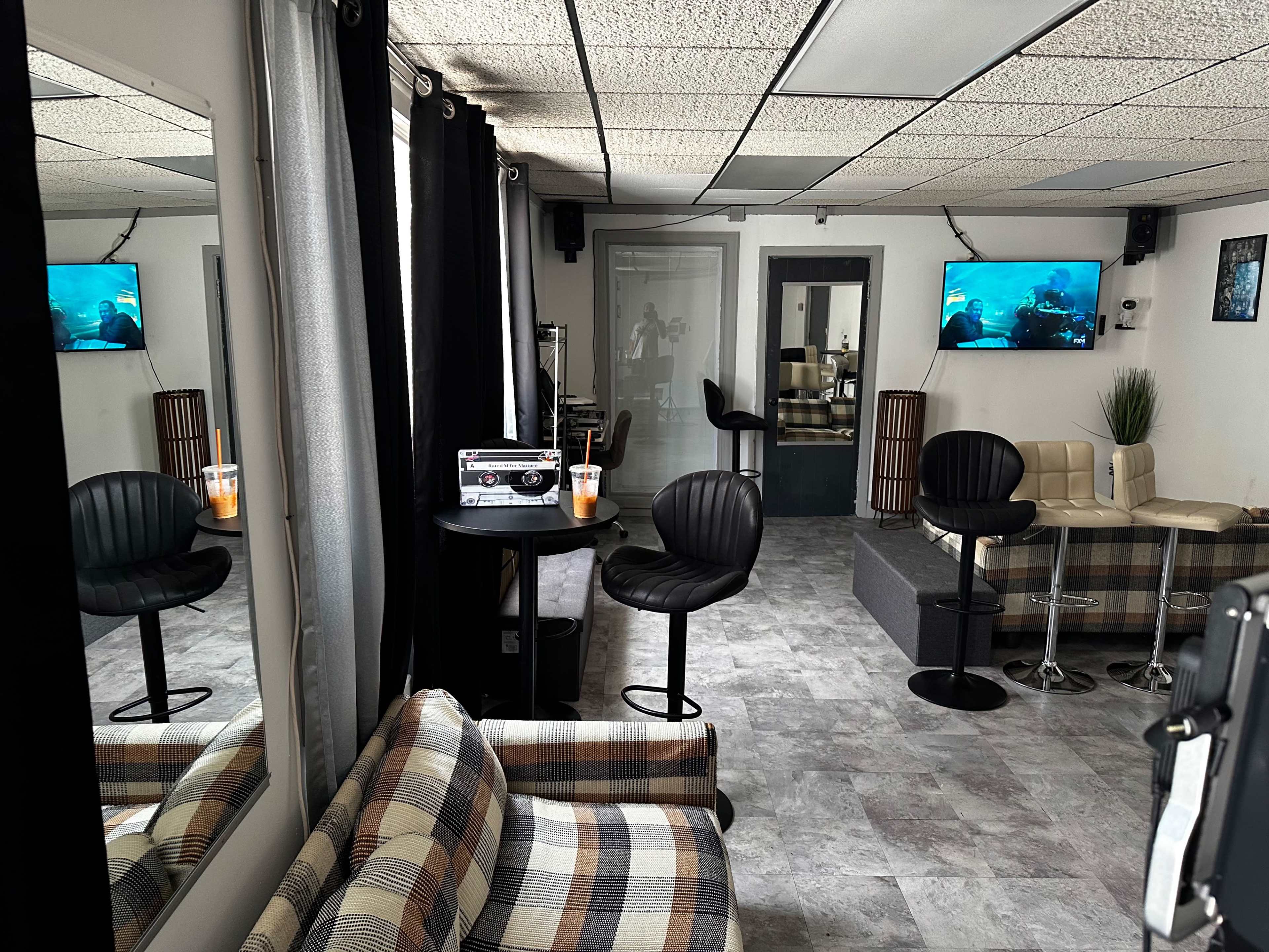 A modern lounge area with a checkered sofa, black bar stools, and two televisions mounted on the walls.