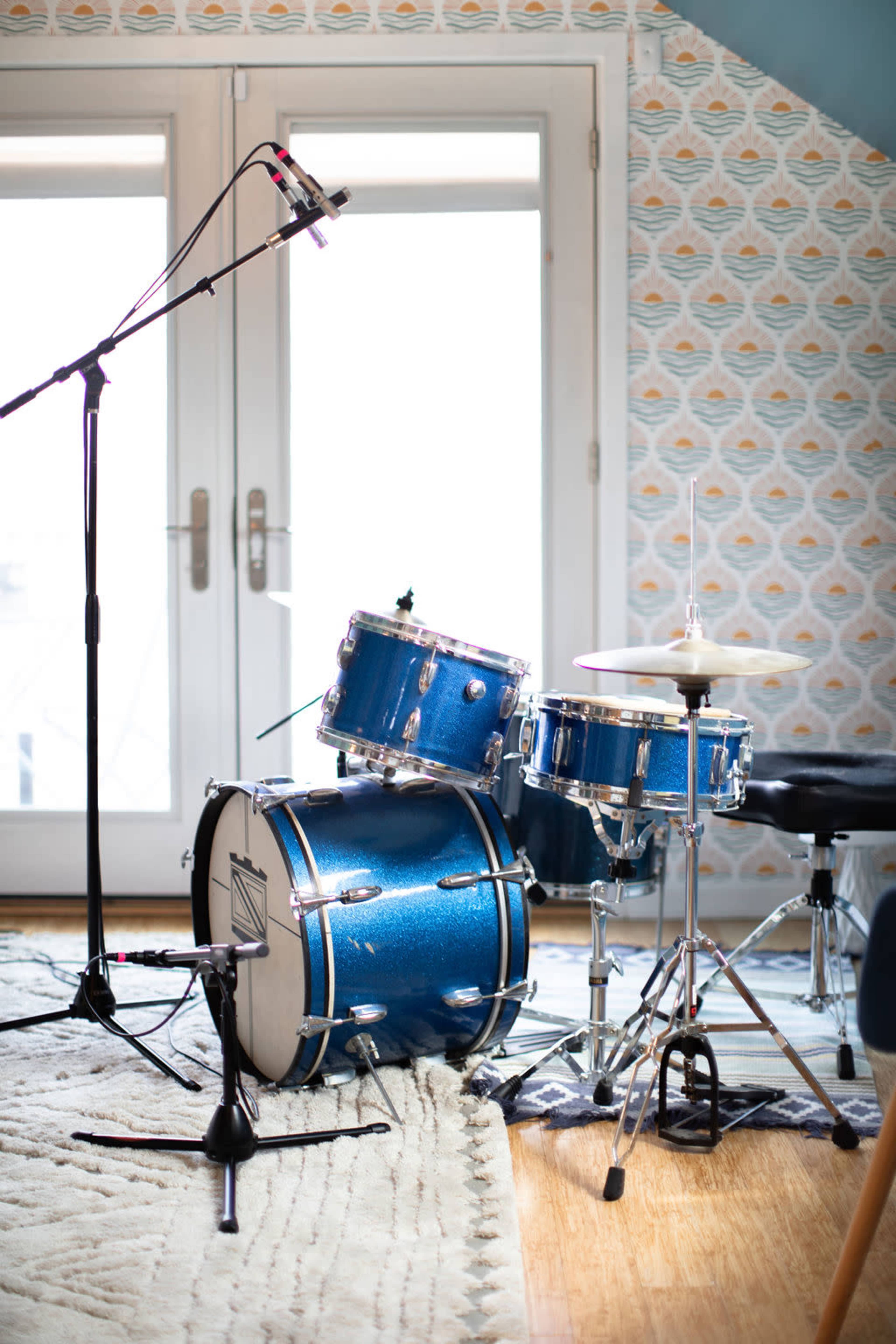 A blue drum set is positioned on a rug in front of a window, accompanied by a microphone on a stand.
