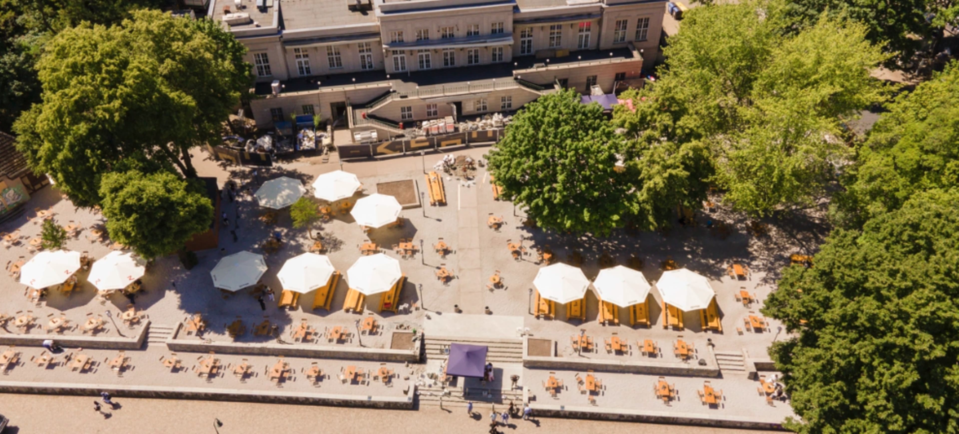An aerial view of a sunny outdoor seating area with umbrellas and tables, surrounded by trees and a building in the background.