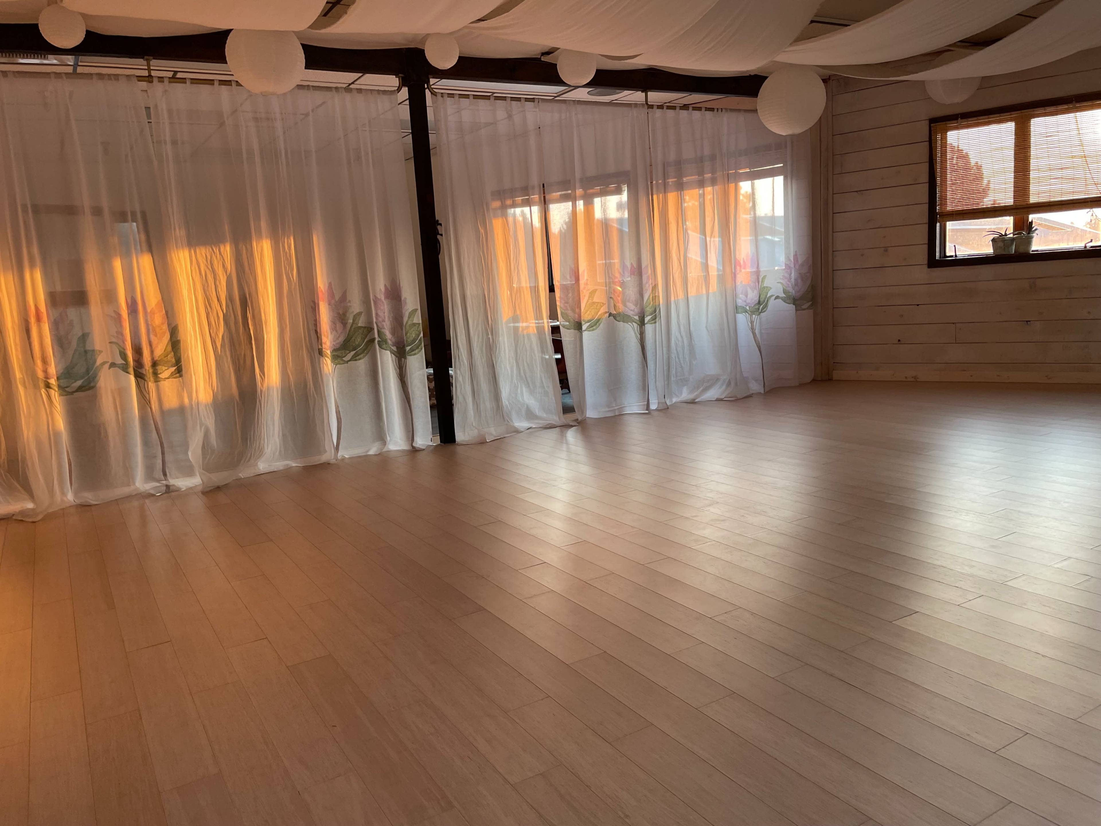 A spacious, light-filled room with wooden floors, sheer curtains featuring floral designs, and large windows allowing sunlight to stream in.