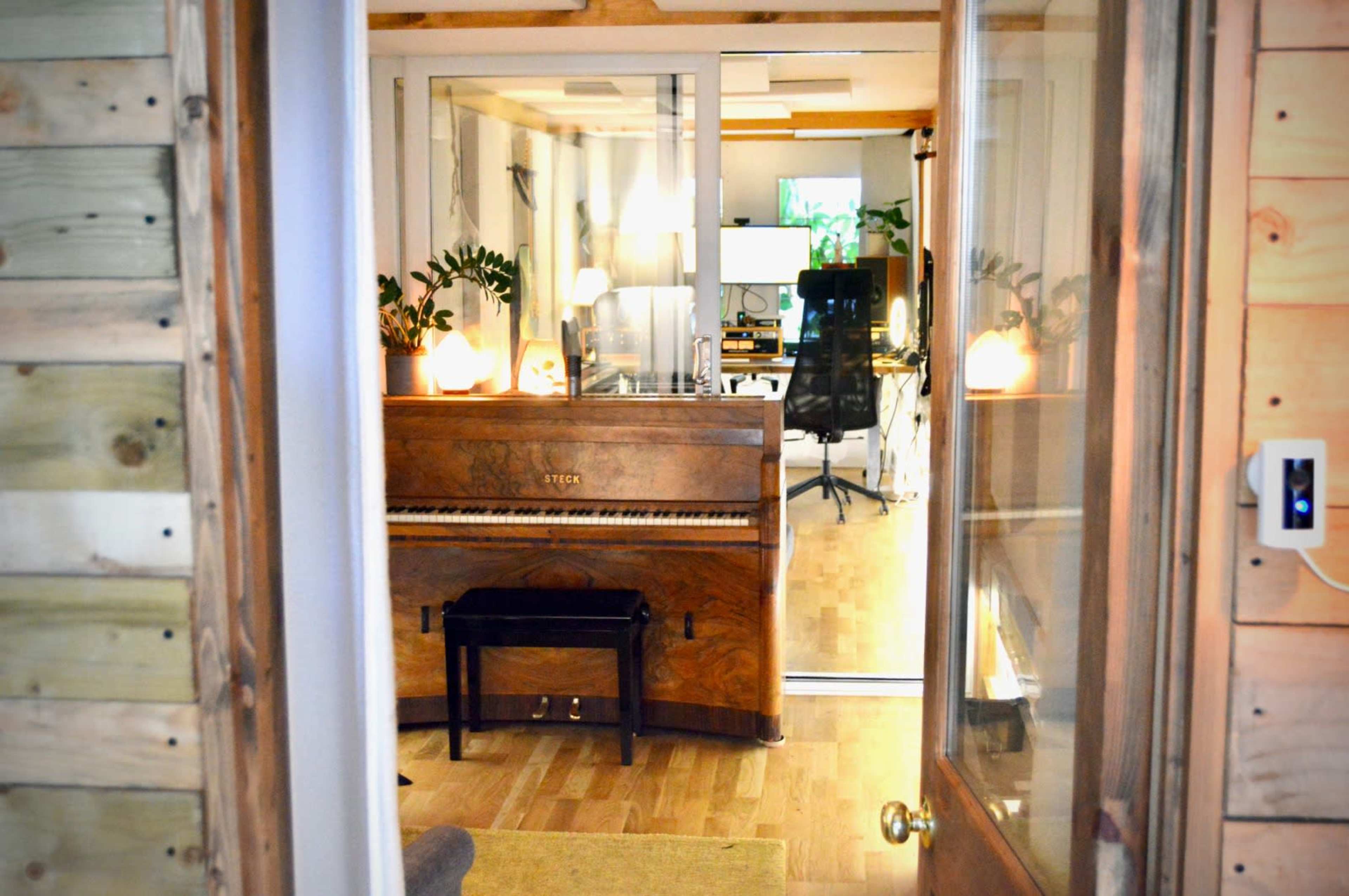 A wooden piano sits in a softly lit room, visible through a glass door that leads to a workspace with computers and plants.