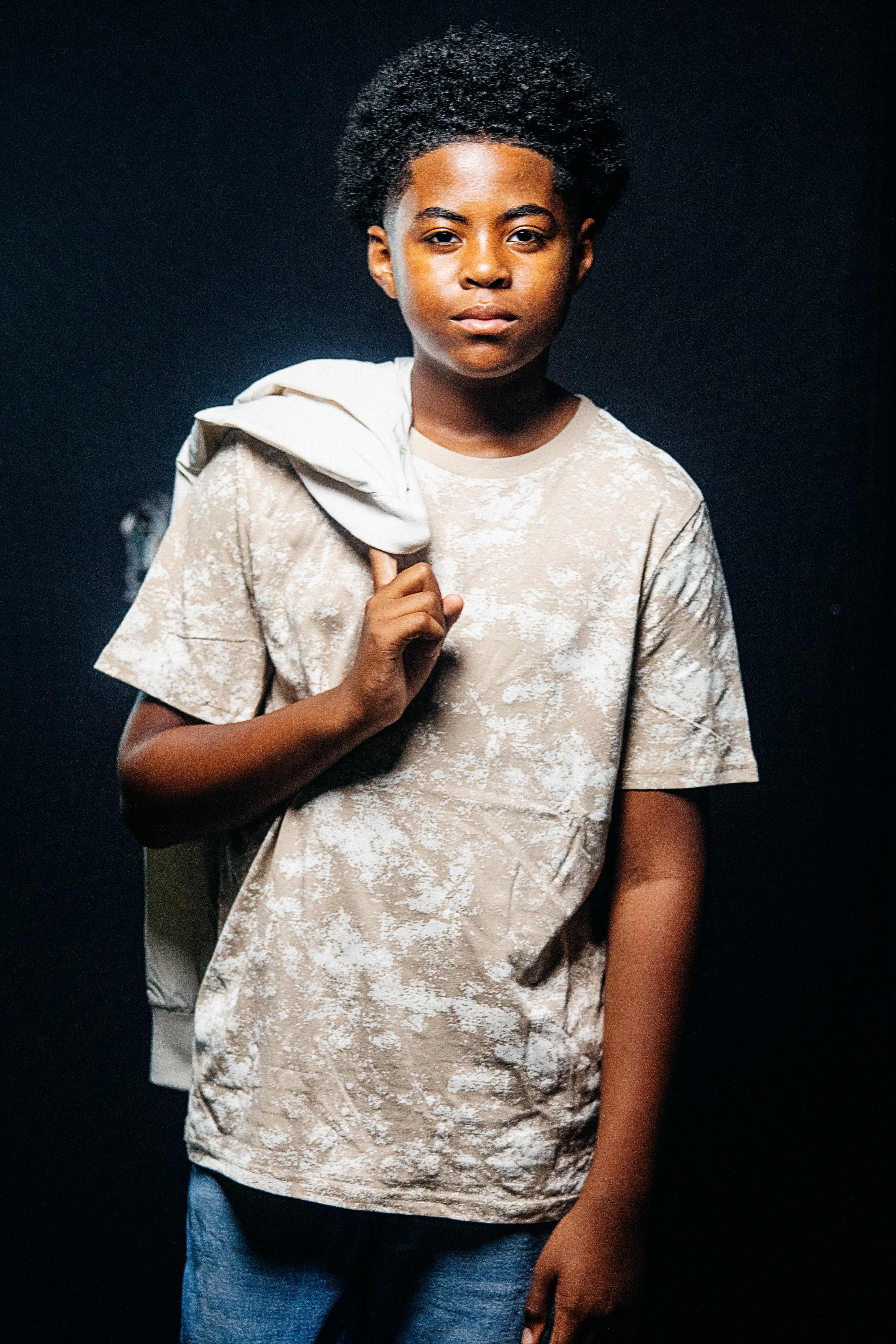 A young person stands against a dark background, wearing a beige patterned shirt and holding a light-colored jacket over their shoulder.