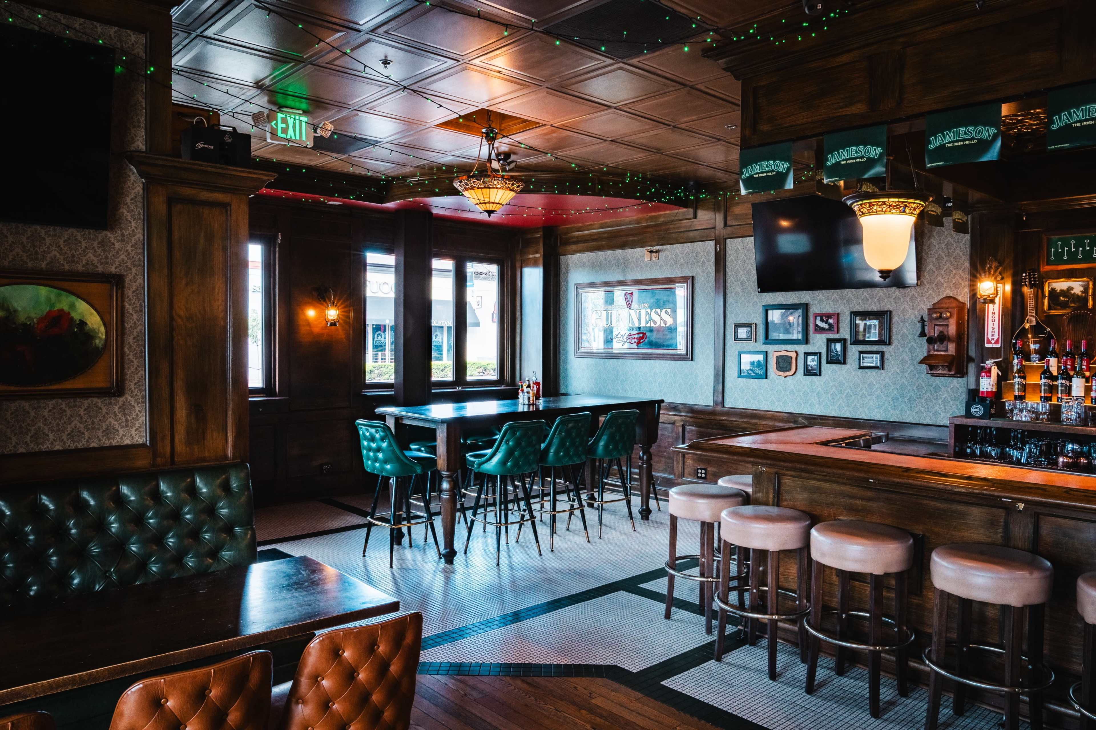 The image shows the interior of a bar with wooden furnishings, green upholstered chairs, and several tables arranged around a bar area.