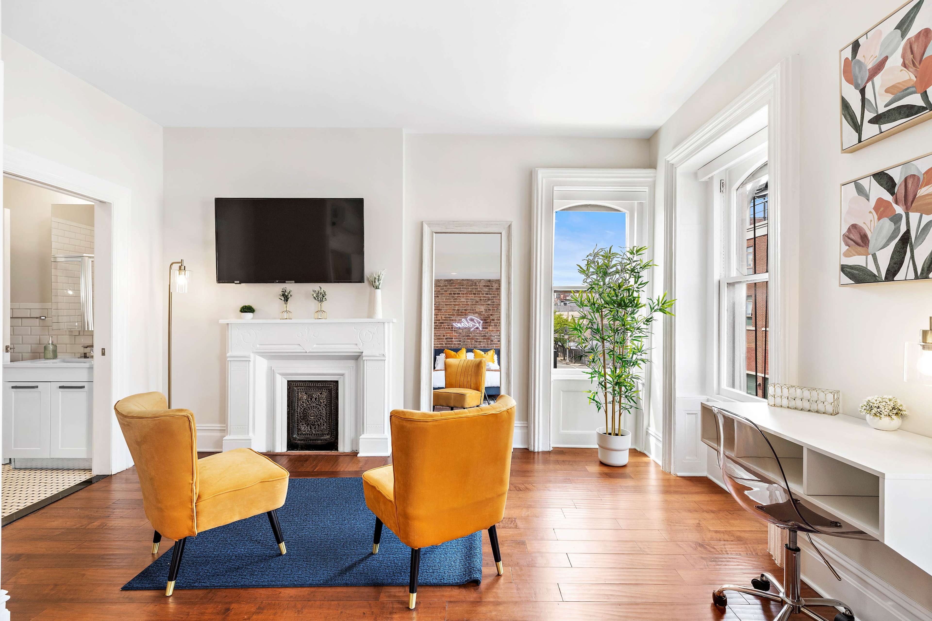 A bright room featuring two yellow chairs facing a white fireplace, a flat-screen TV mounted on the wall, and a desk near a window.