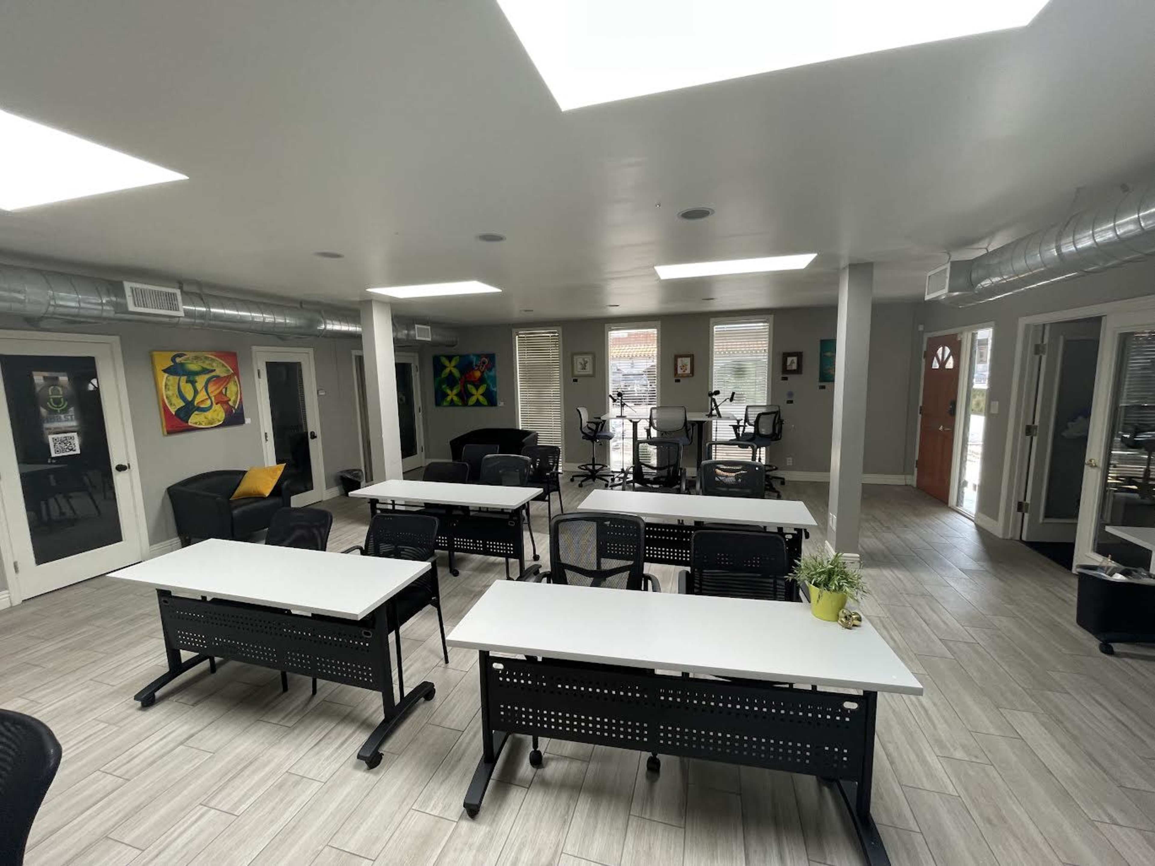 A spacious, well-lit room with multiple desks arranged in rows, surrounded by chairs and artwork on the walls.