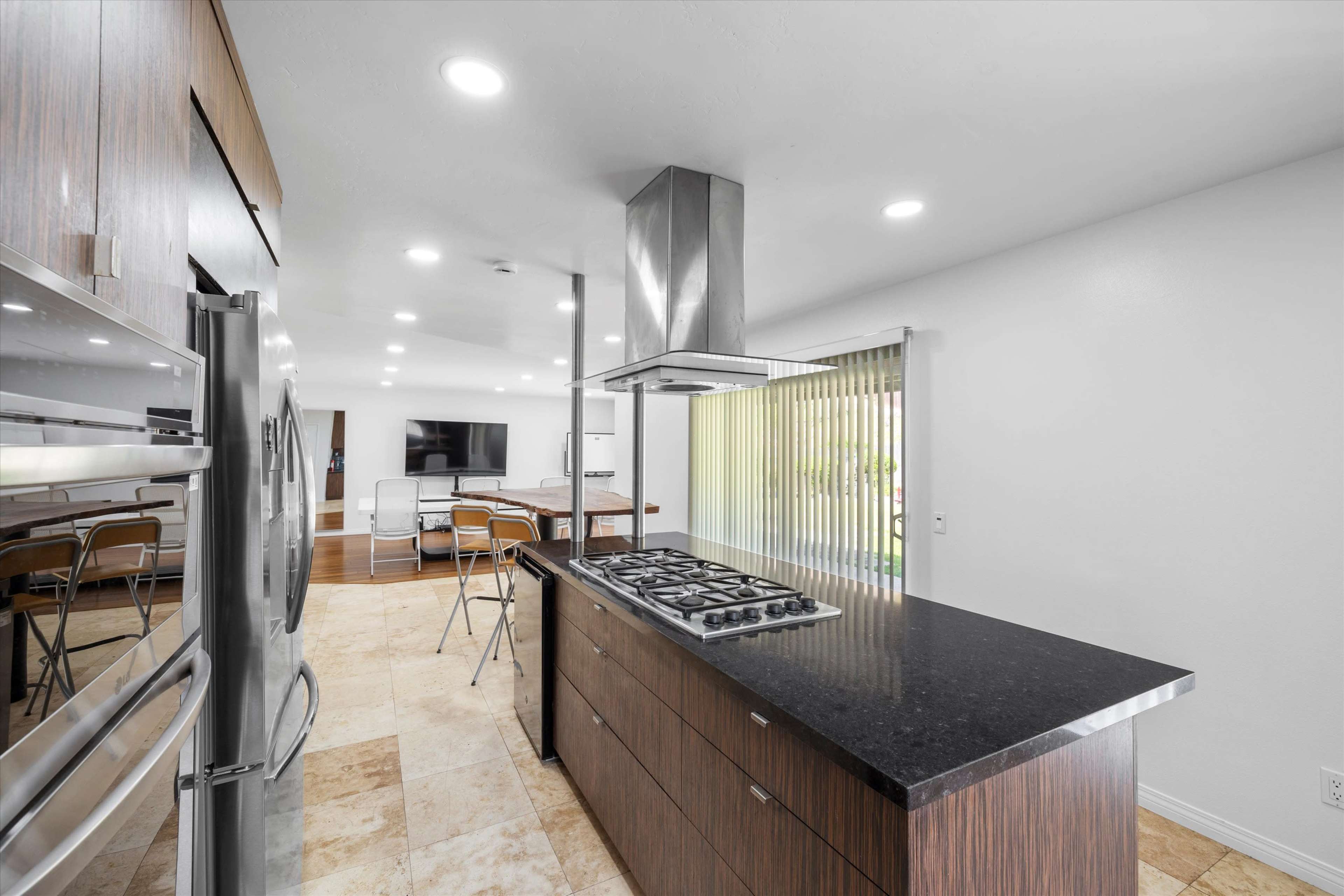 The image shows a modern kitchen featuring a central island with a gas cooktop, stainless steel appliances, and a dining area visible in the background.