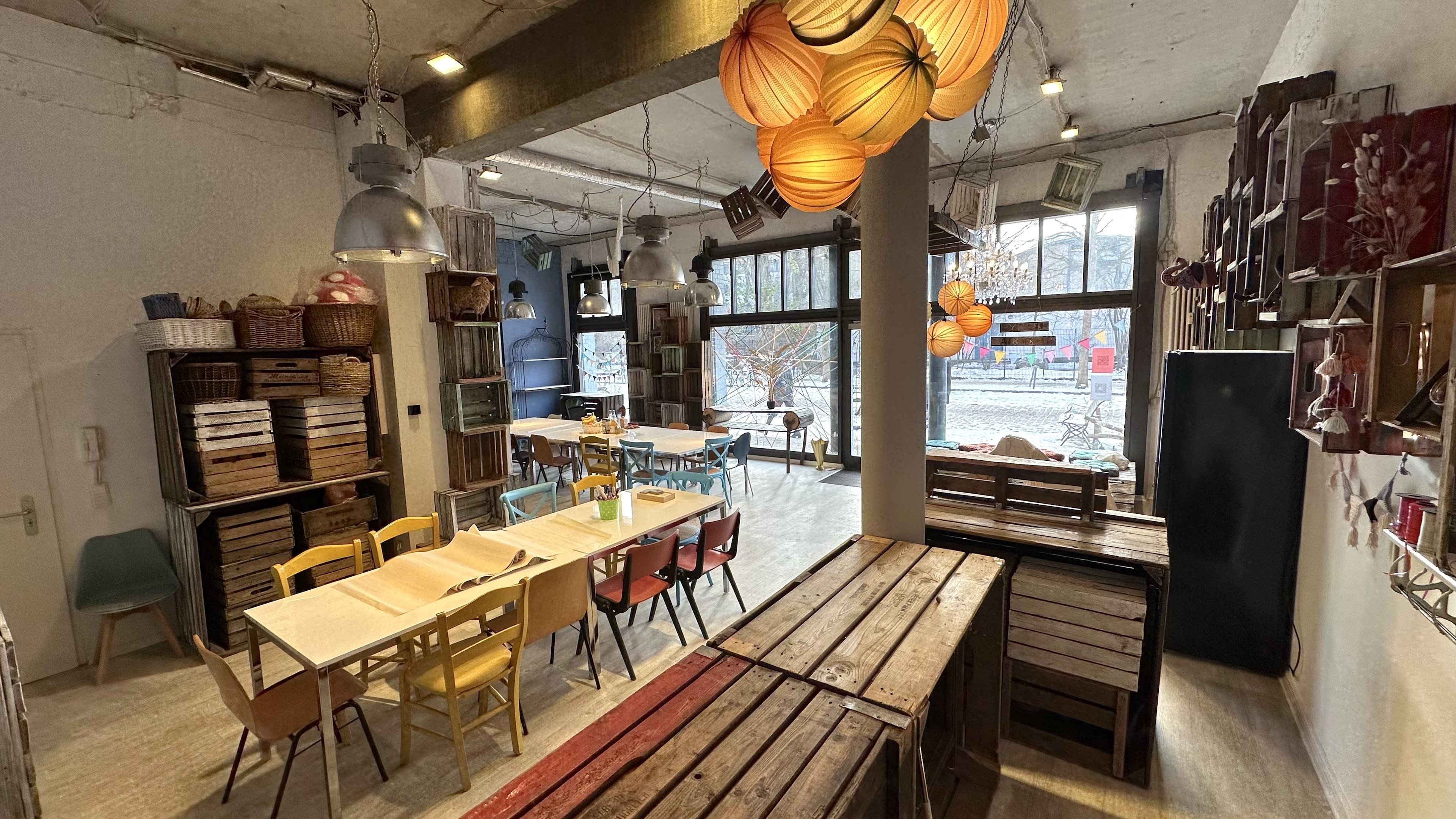 The image shows a cozy café interior with wooden crates as furniture, colorful chairs, and decorative lighting hanging from the ceiling.
