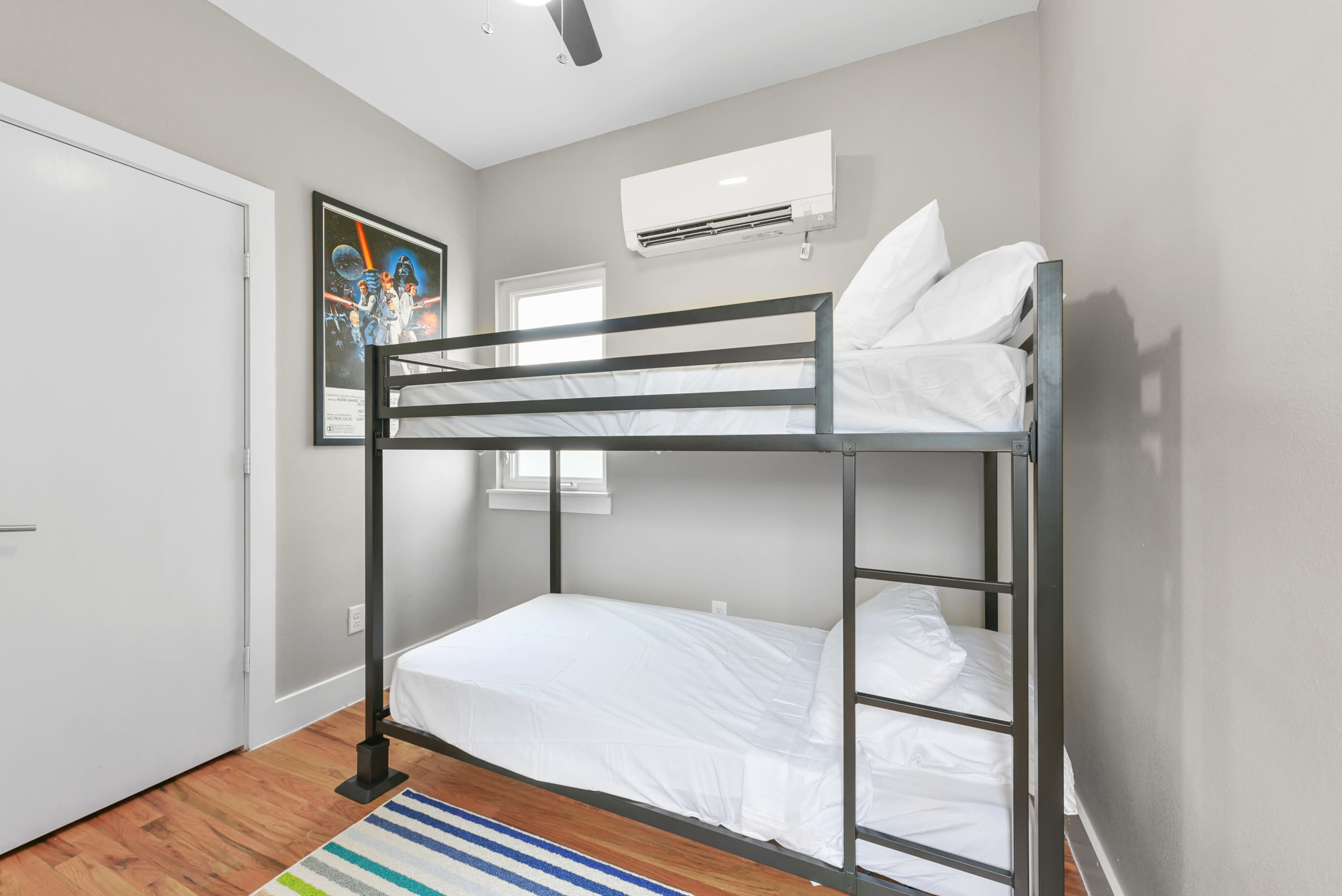 The image shows a small room featuring a metal bunk bed with unmade white bedding, a window, and an air conditioning unit on the wall.