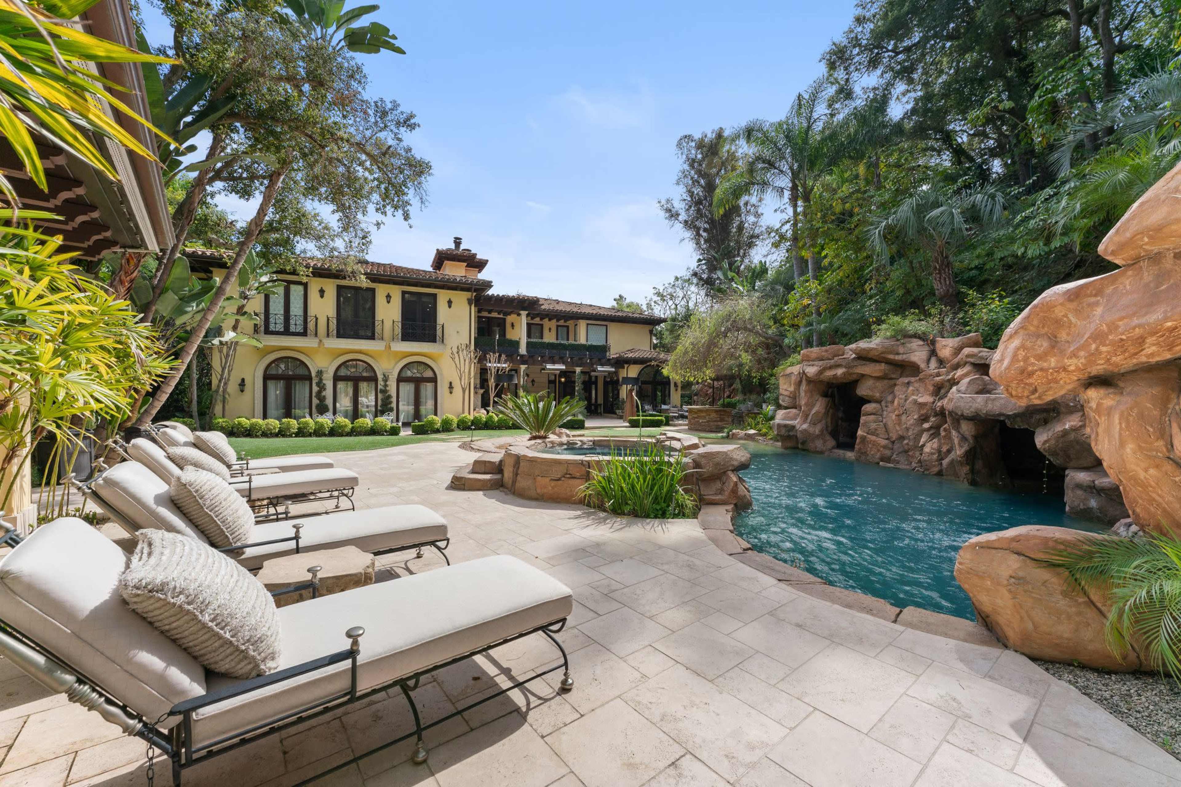 A luxurious backyard features a swimming pool with rock formations, lounging chairs, and a large property surrounded by greenery.