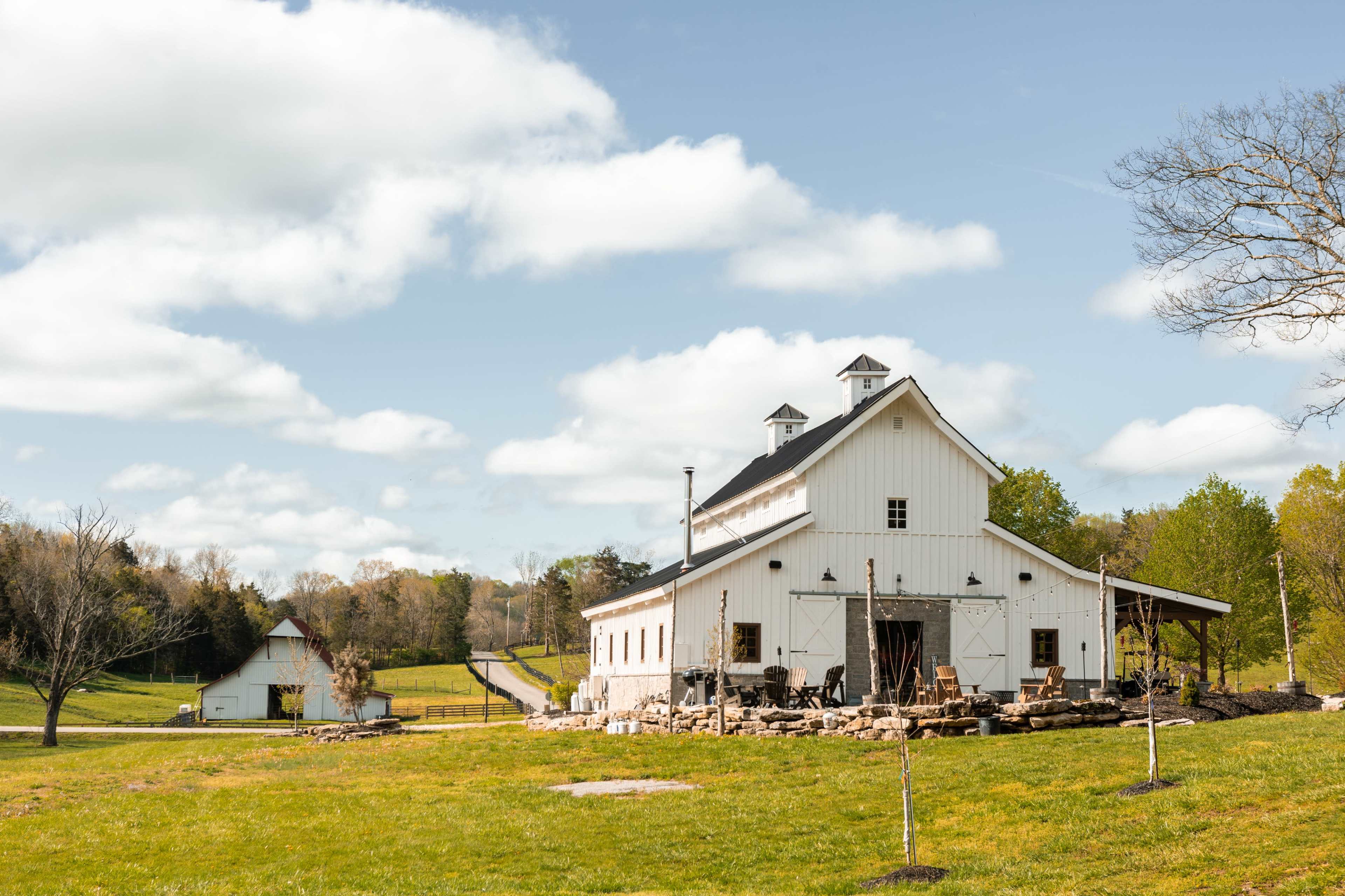 A white barn with a stone patio and nearby trees is set against a cloudy sky in a rural landscape.