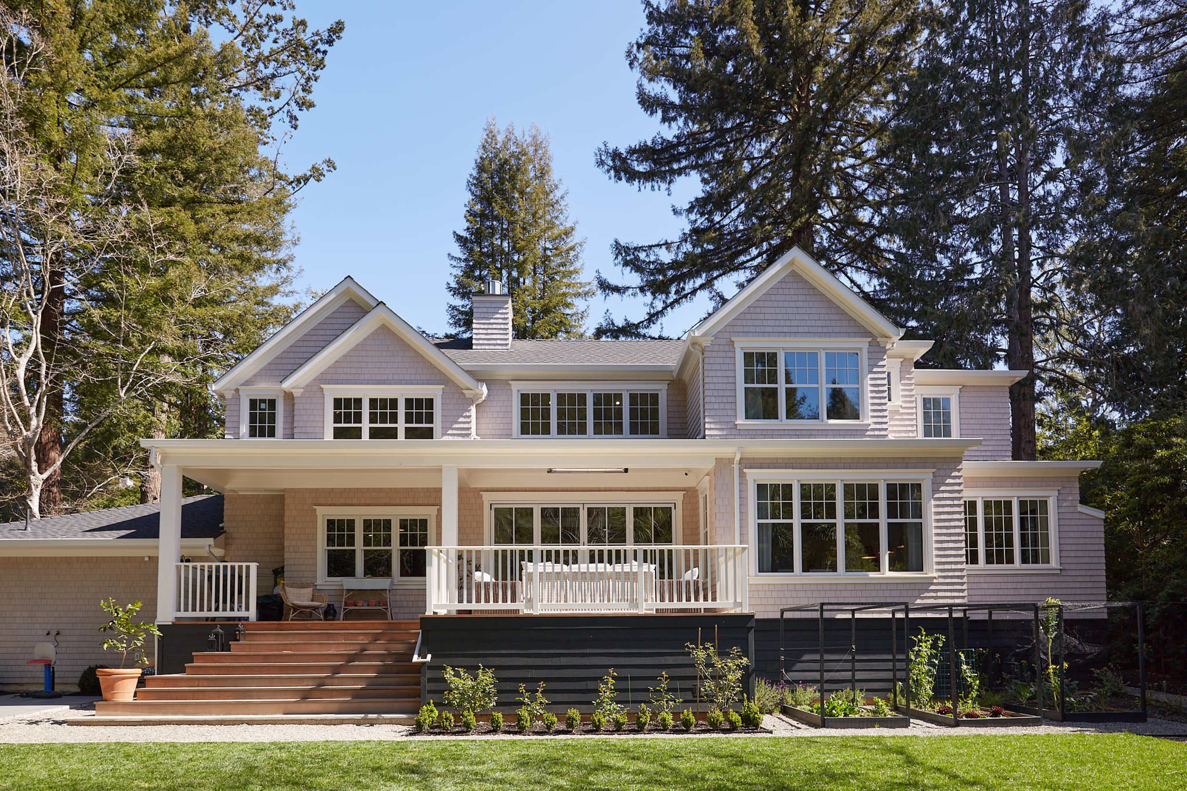 A large, light-colored house features multiple windows, a spacious deck with steps, and is surrounded by tall trees and a green lawn.