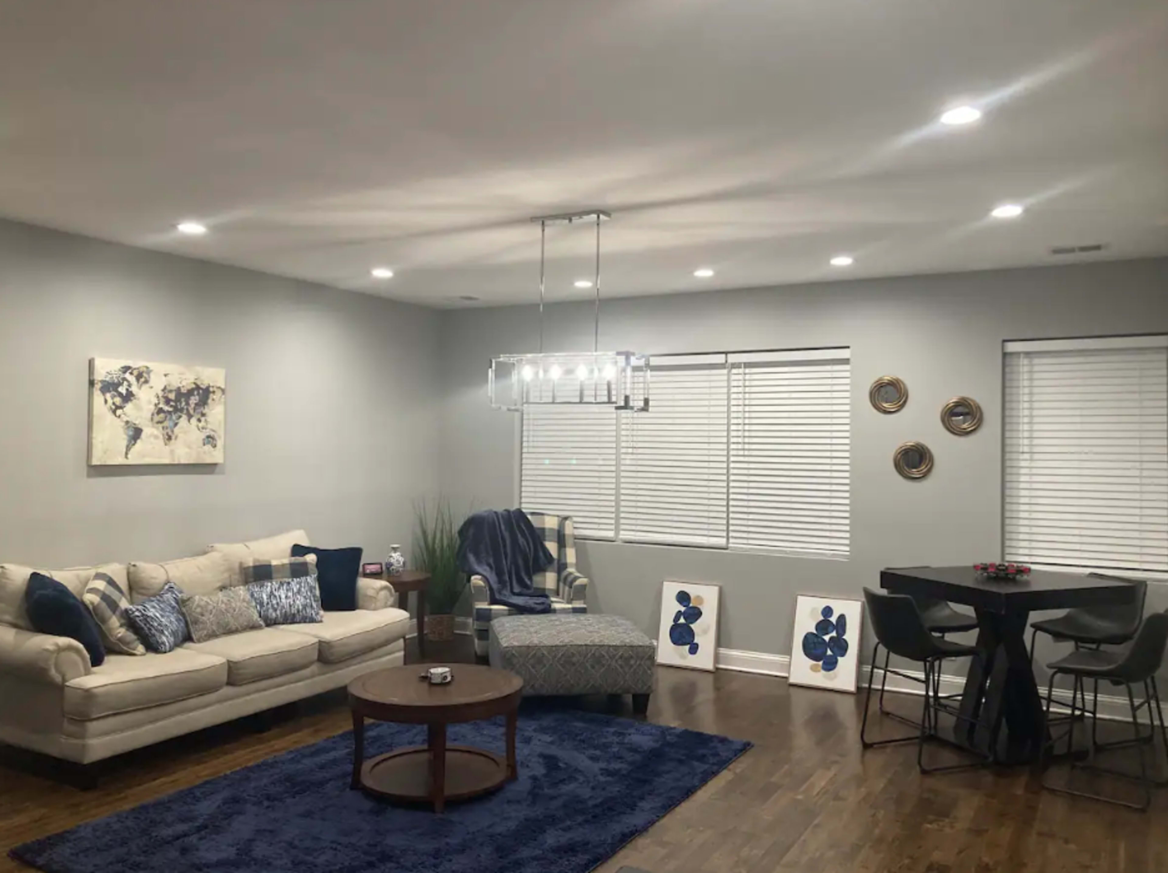 A living room with a beige couch, a round wooden coffee table, a dining area with dark chairs, and large windows adorned with white blinds.