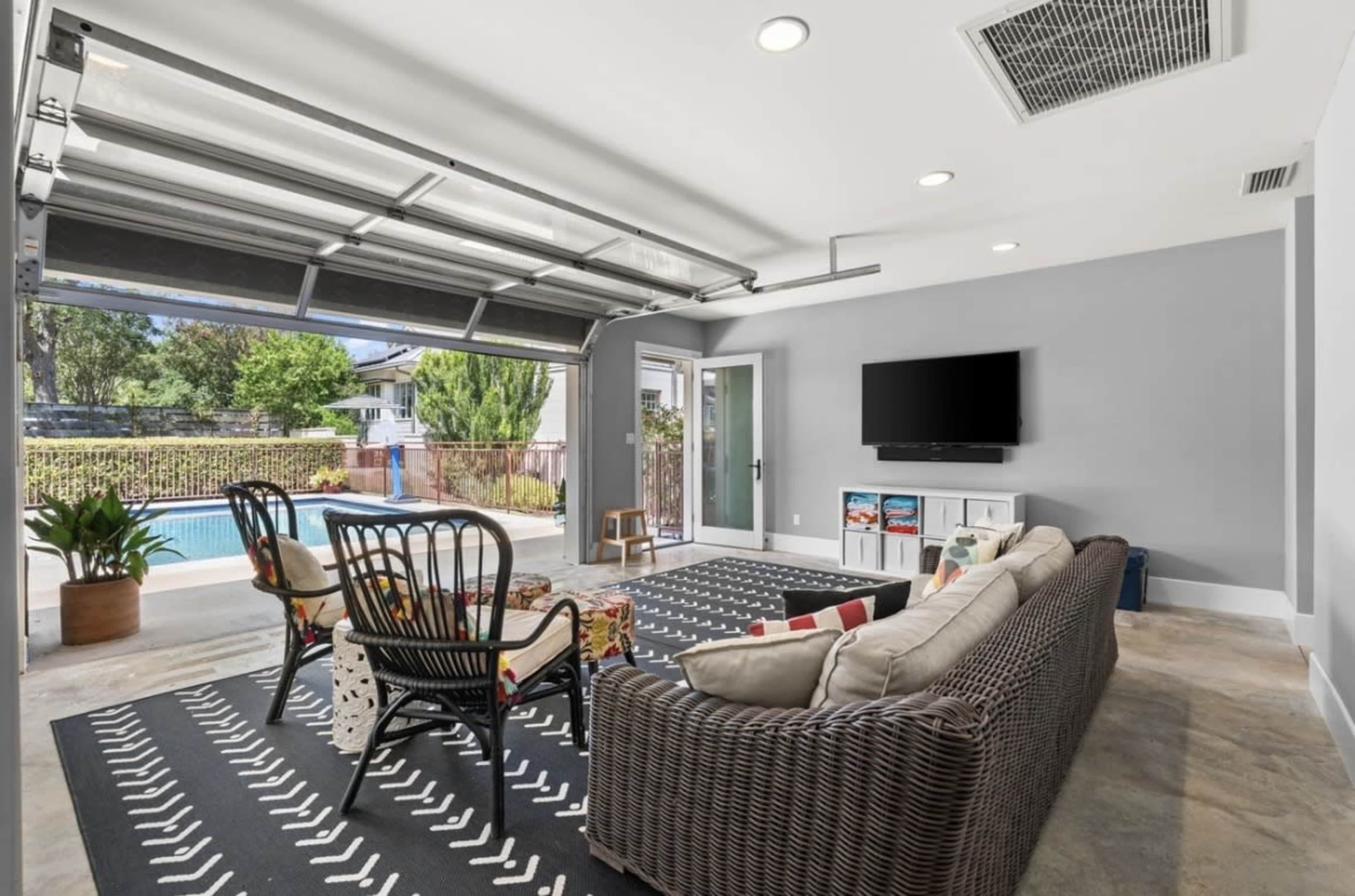 The image shows a modern indoor lounge area with a large open garage door leading to a poolside view, featuring a couch, chairs, a television, and decorative elements.