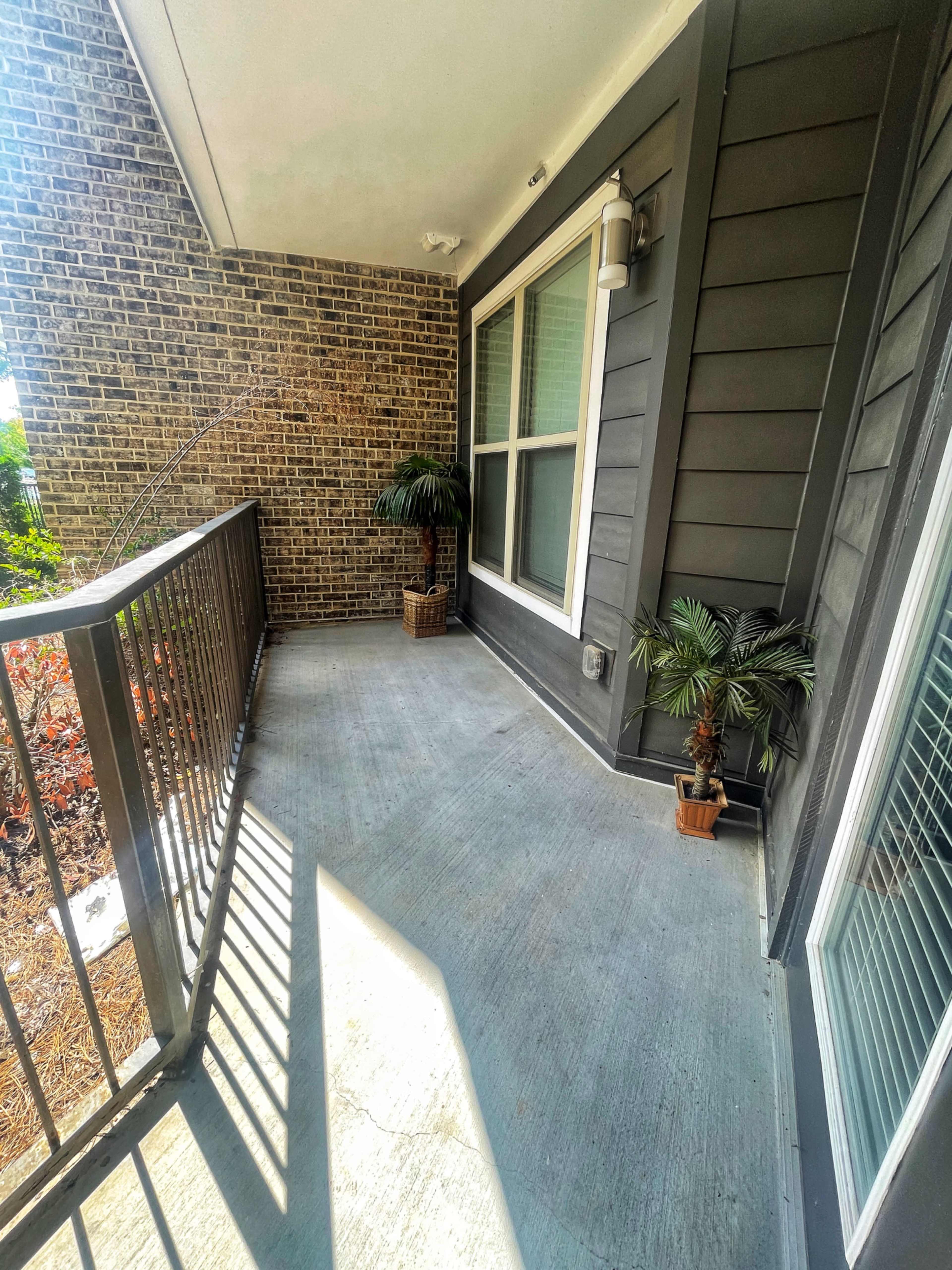 The image shows a small covered balcony with potted plants and a railing, adjacent to a brick wall.