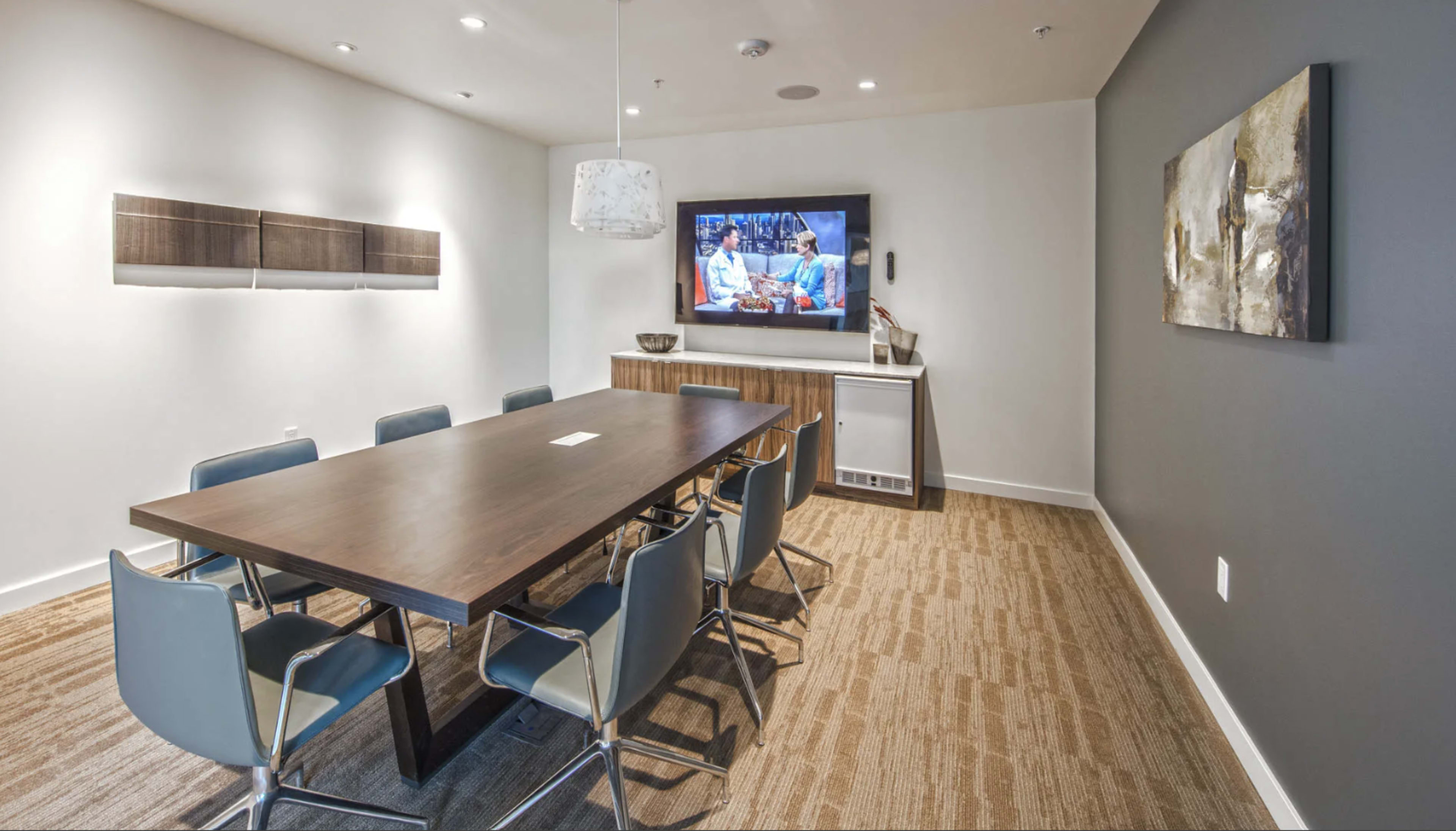 The image shows a modern conference room with a large wooden table surrounded by chairs, a wall-mounted television, and contemporary artwork.