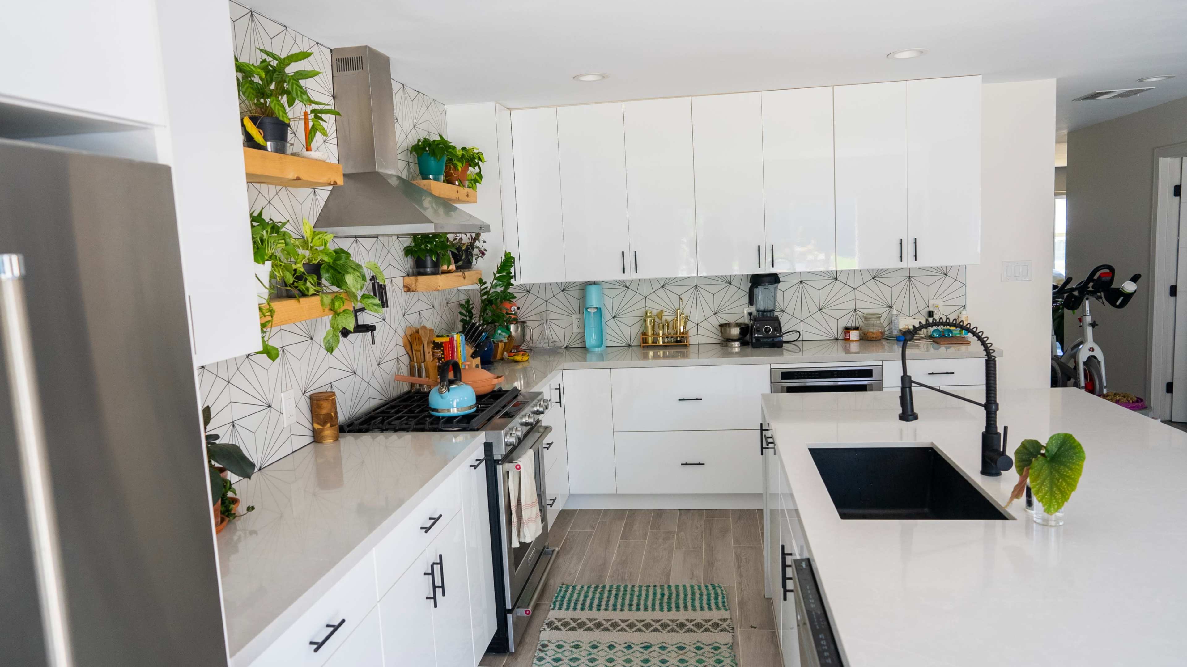 A modern kitchen features white cabinets, a black sink, a gas stove, and shelves adorned with potted plants and decorative items against a geometric tiled backsplash.