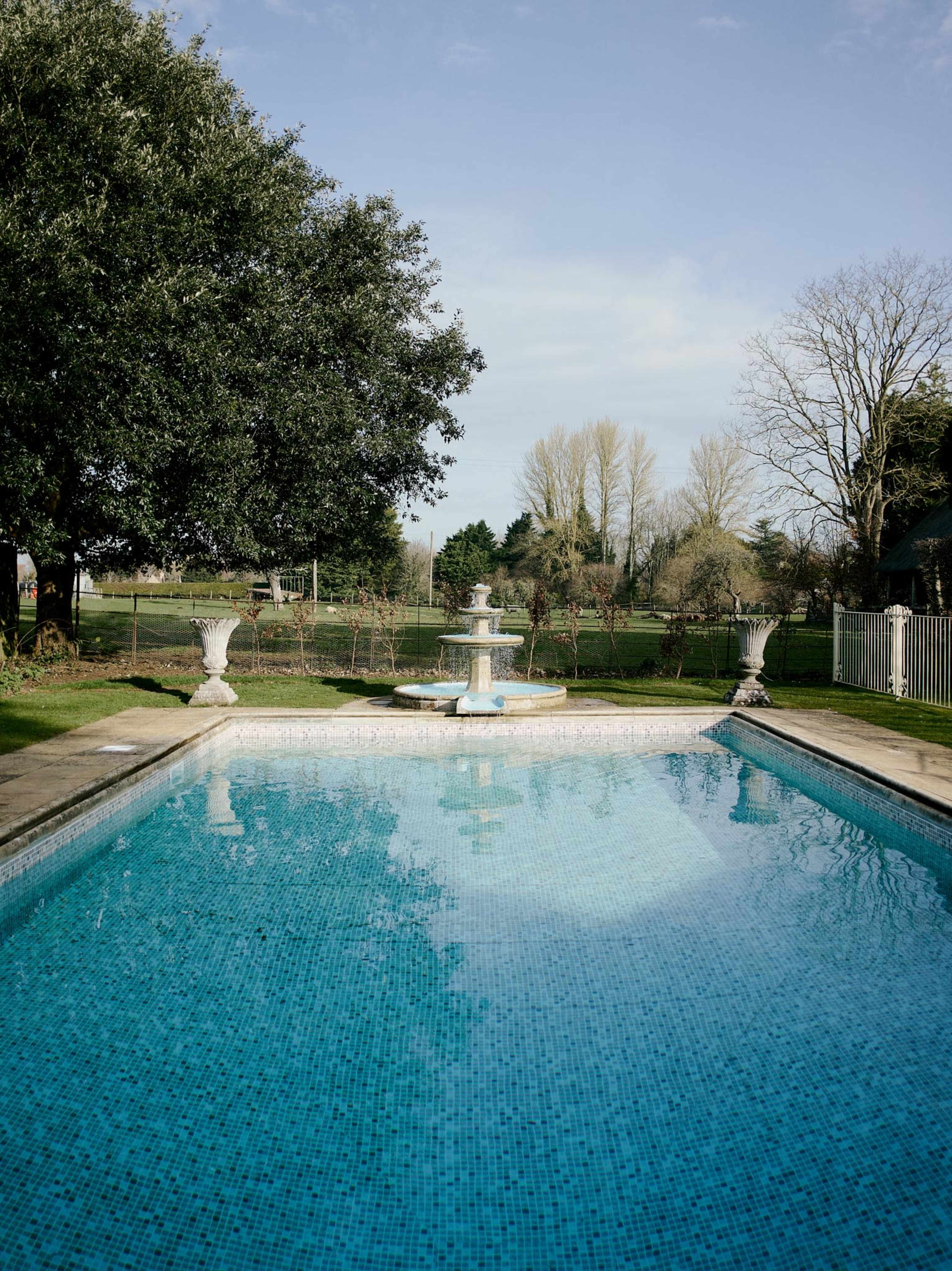A clear blue swimming pool features a fountain at its center, surrounded by greenery and a fence in the background.