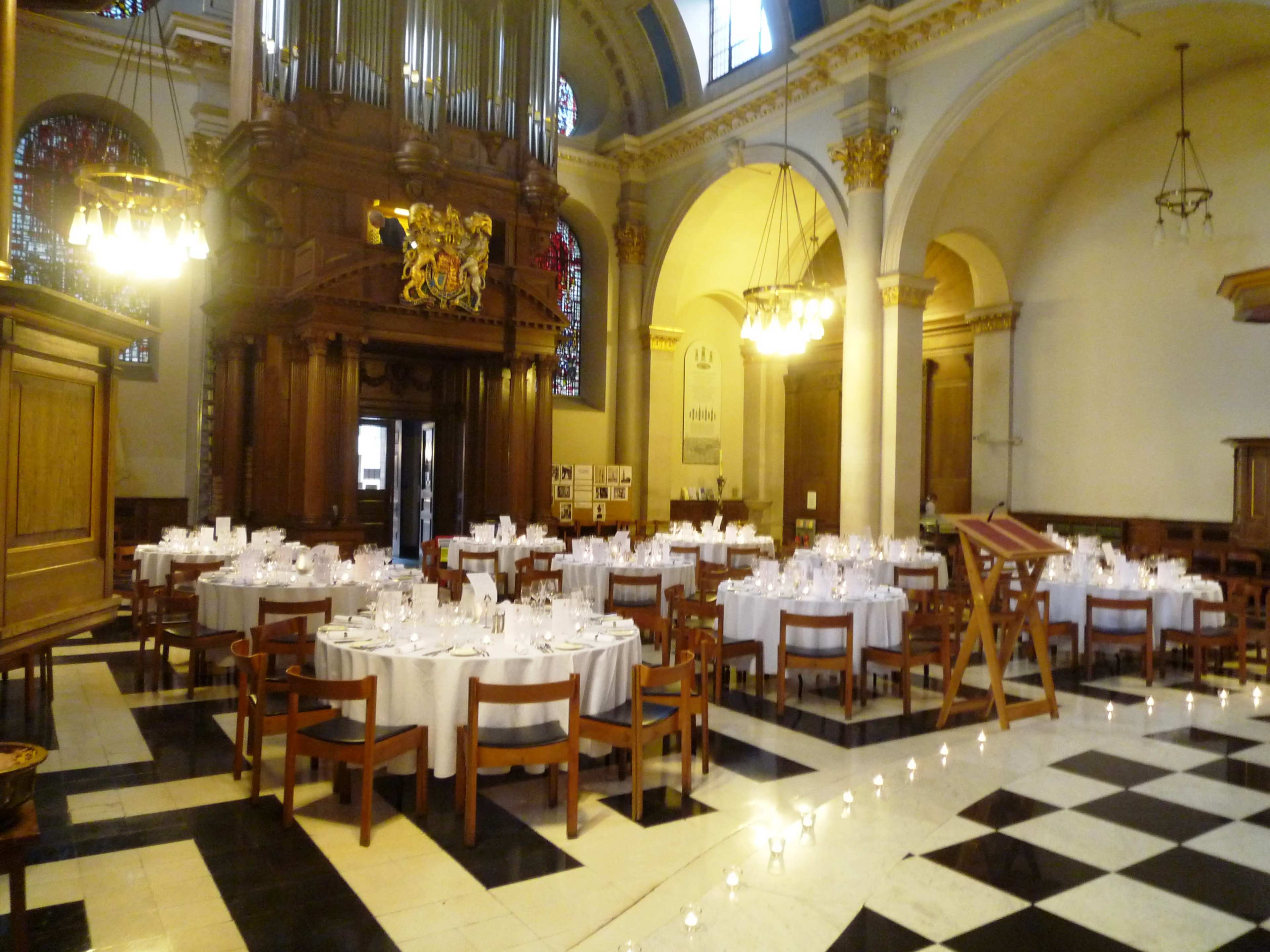 The image shows a banquet setup with round tables covered in white tablecloths arranged in a large hall featuring high ceilings and ornate architectural details.
