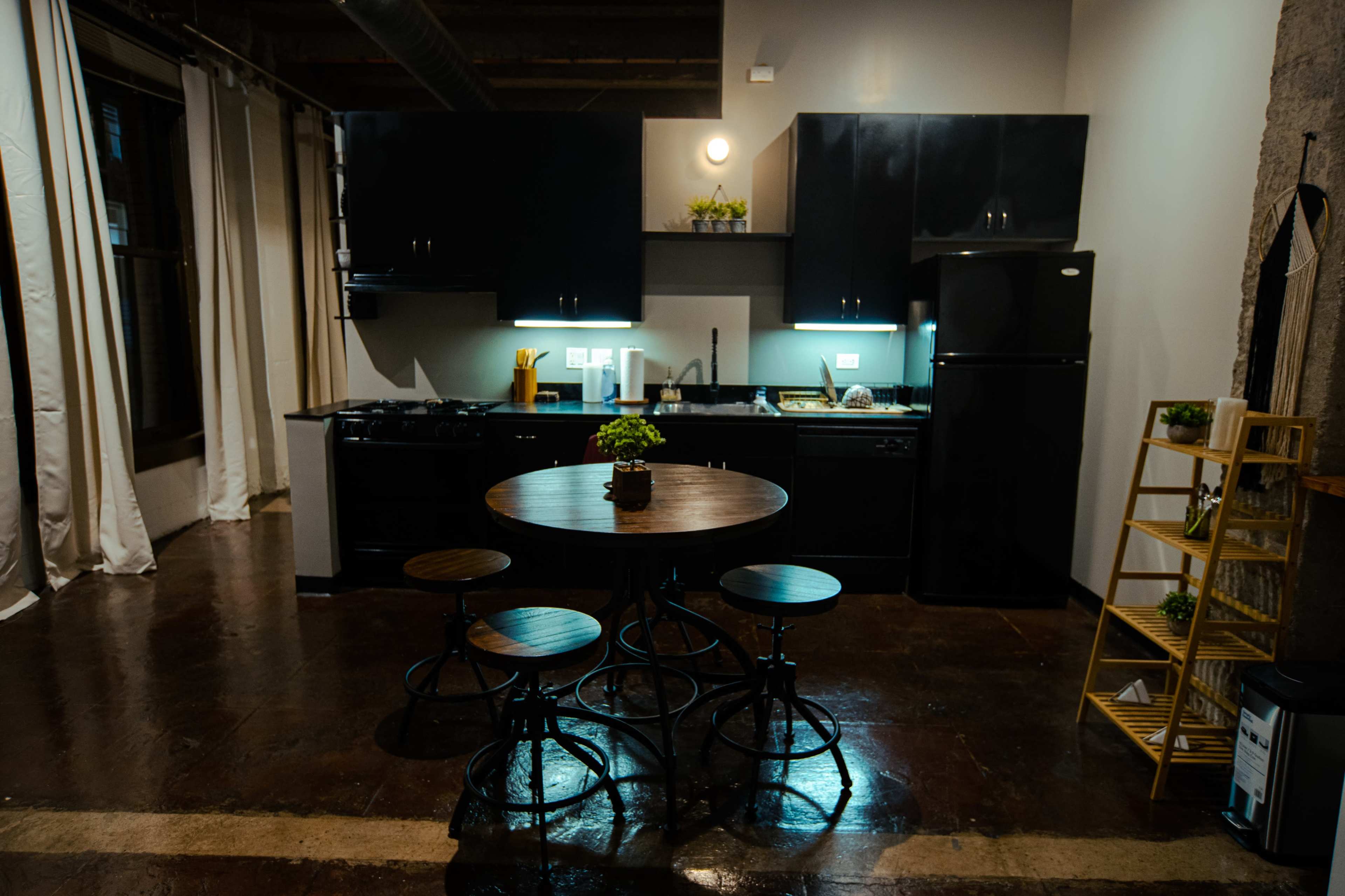A modern kitchen with black cabinets, a circular wooden dining table, and three stools is illuminated by under-cabinet lighting.