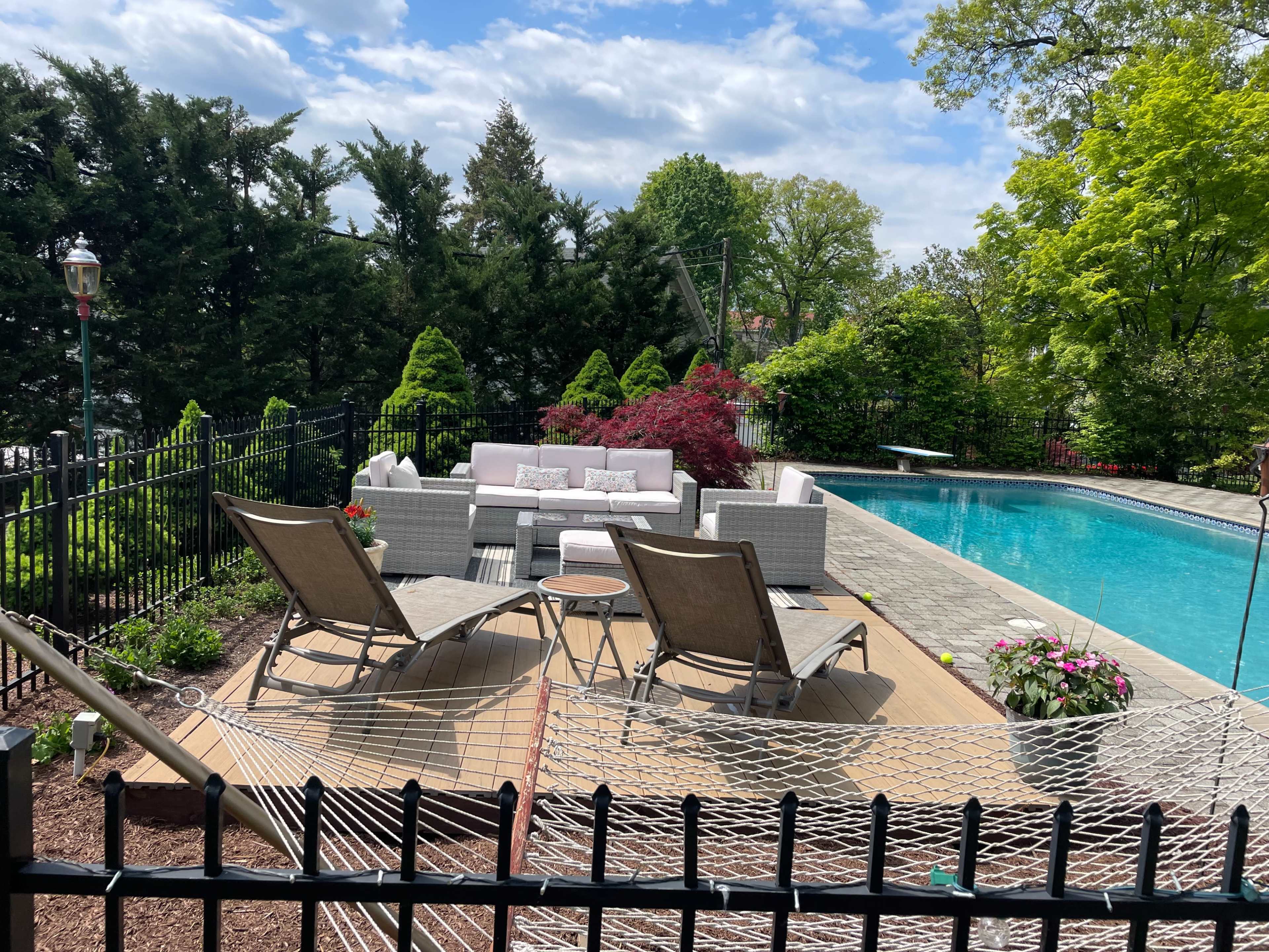 The image shows a landscaped backyard with a swimming pool, lounge chairs, a hammock, and a seating area surrounded by greenery.
