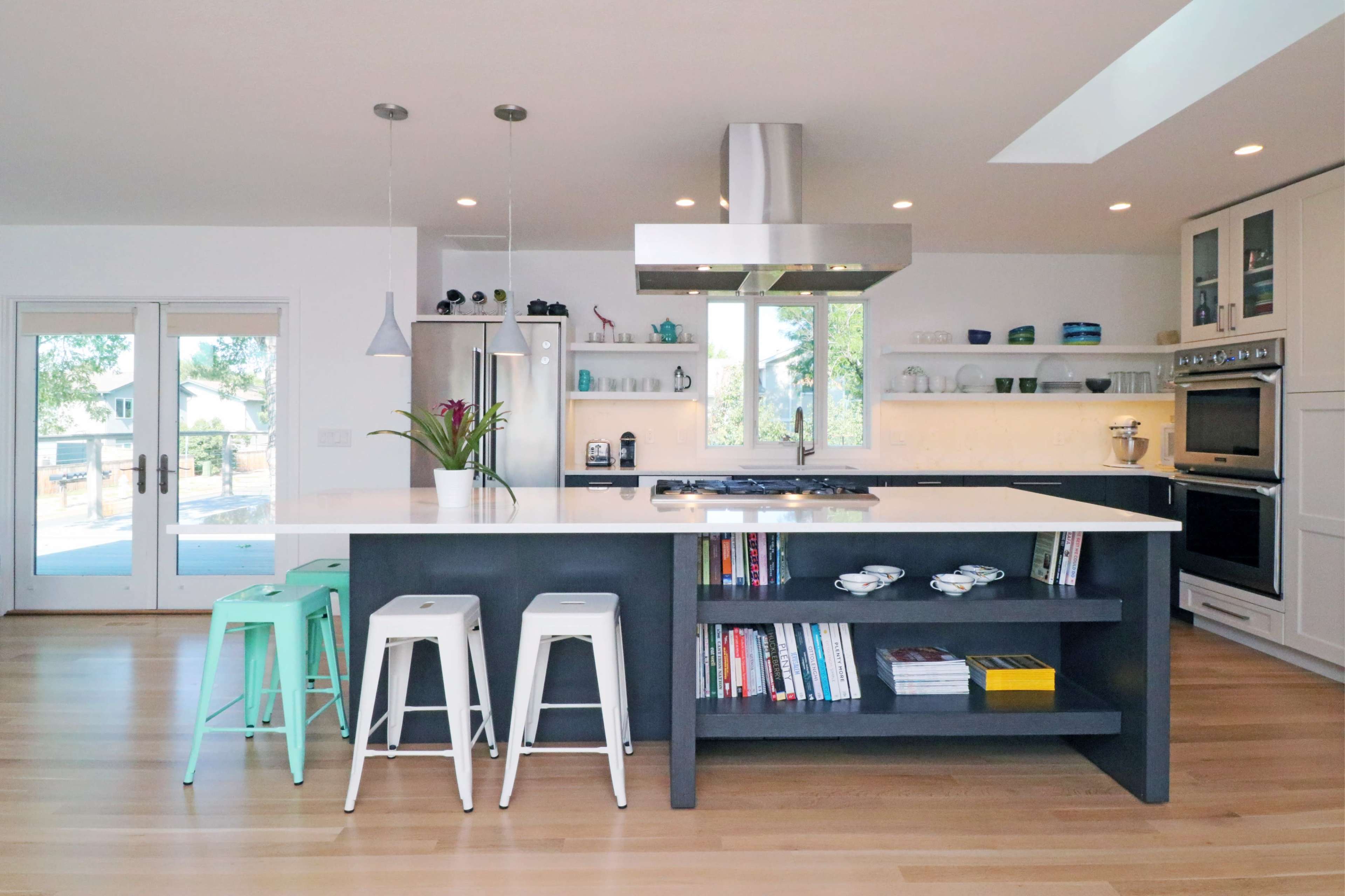 A modern kitchen with a large island, stainless steel appliances, and a light color scheme, featuring bar stools and shelves with books and tableware.