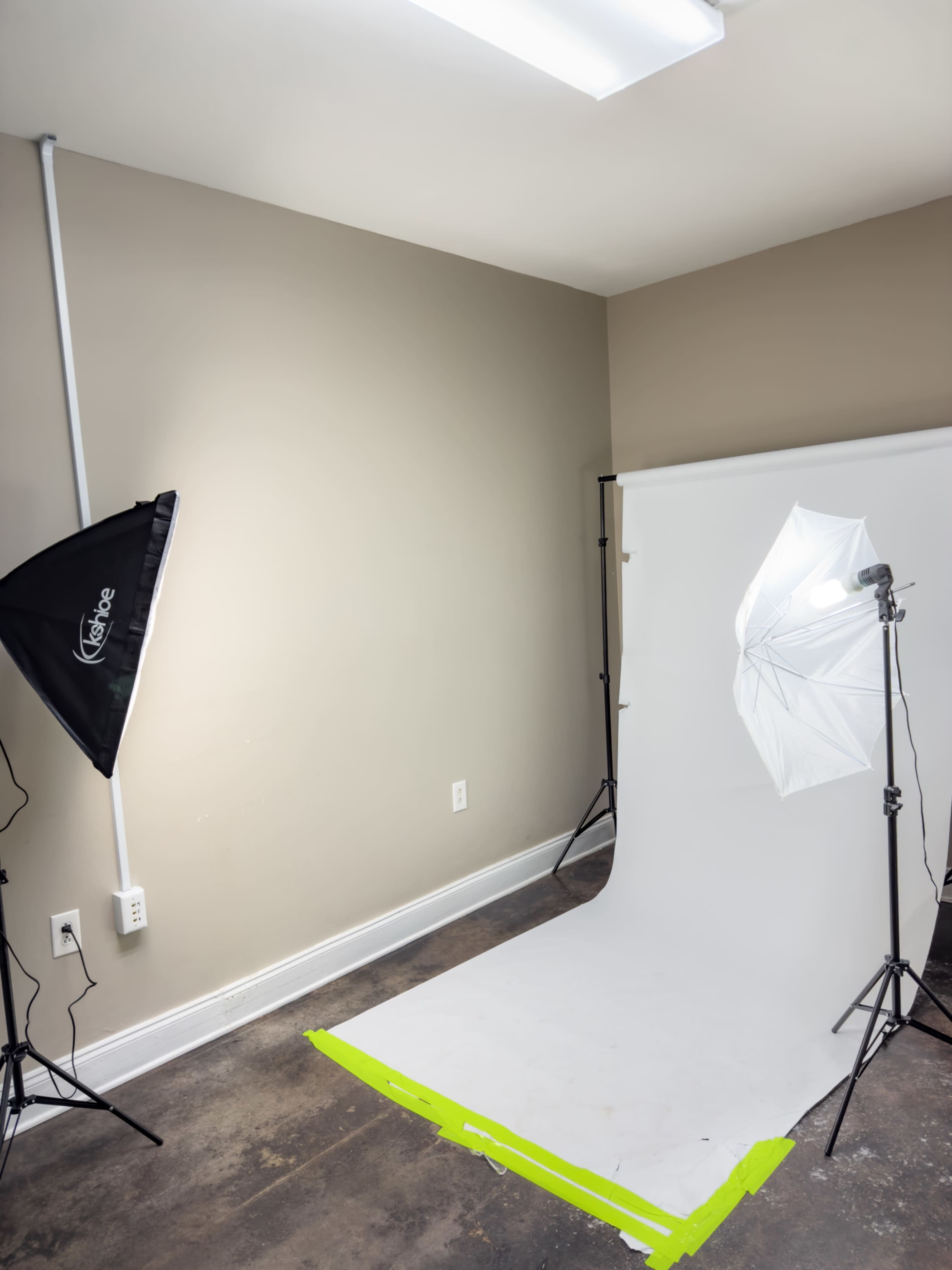The image shows a photography studio setup with a white backdrop, two lighting equipment stands, and a light reflector positioned on the floor.