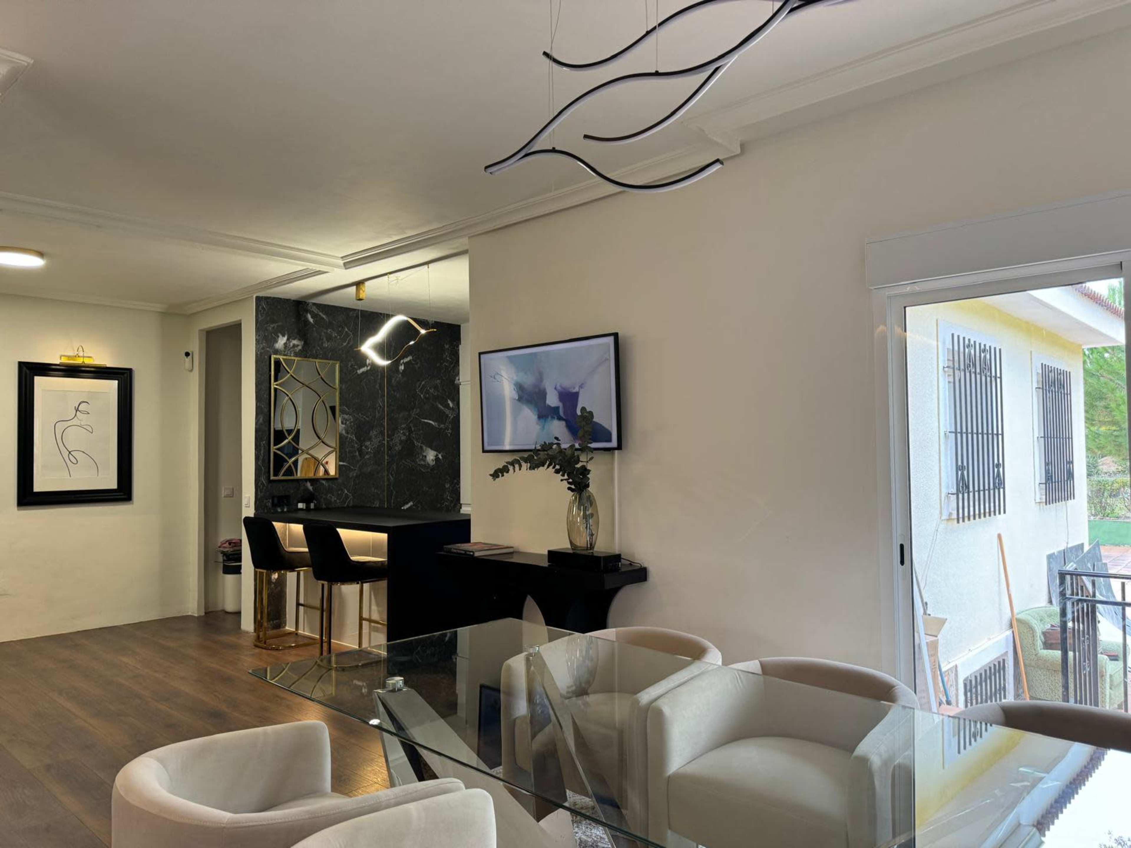 The image shows a modern interior space with a dining area featuring a glass table and beige chairs, adjacent to a stylish bar area with high stools and decorative elements.