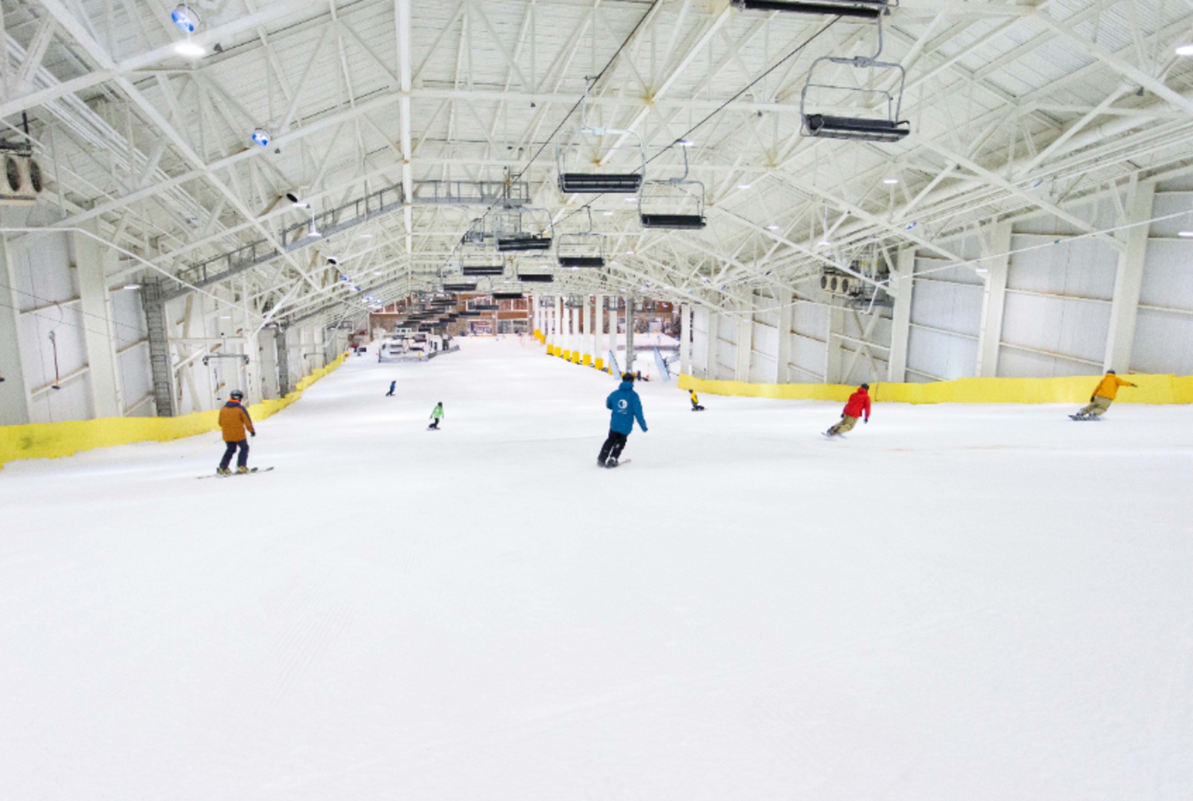 Skiers navigate a wide indoor slope under bright lights in a large, open facility.