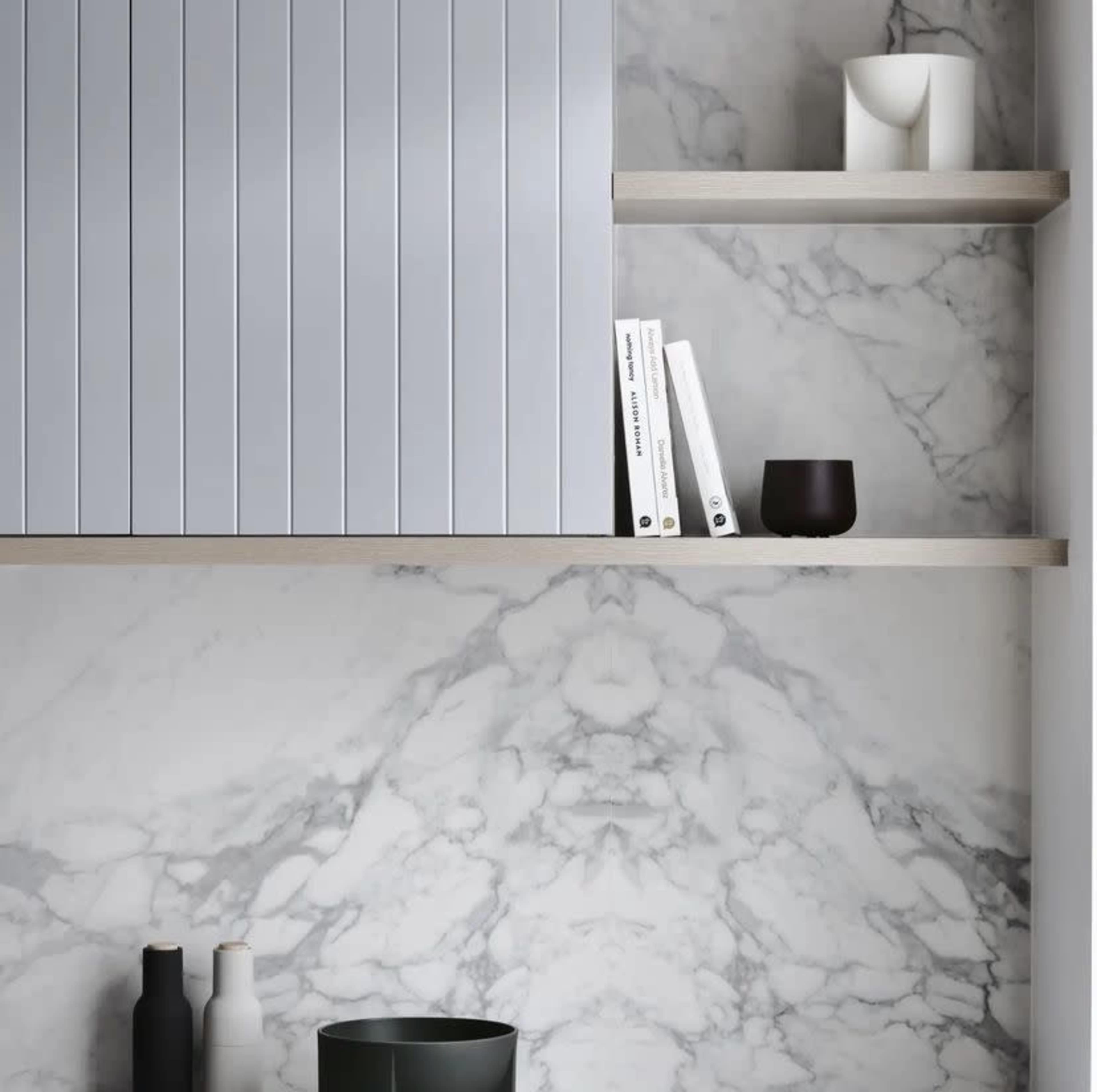 A minimalist interior with gray cabinet doors, wooden shelves, and various decorative items, including bottles and books, against a marble wall.
