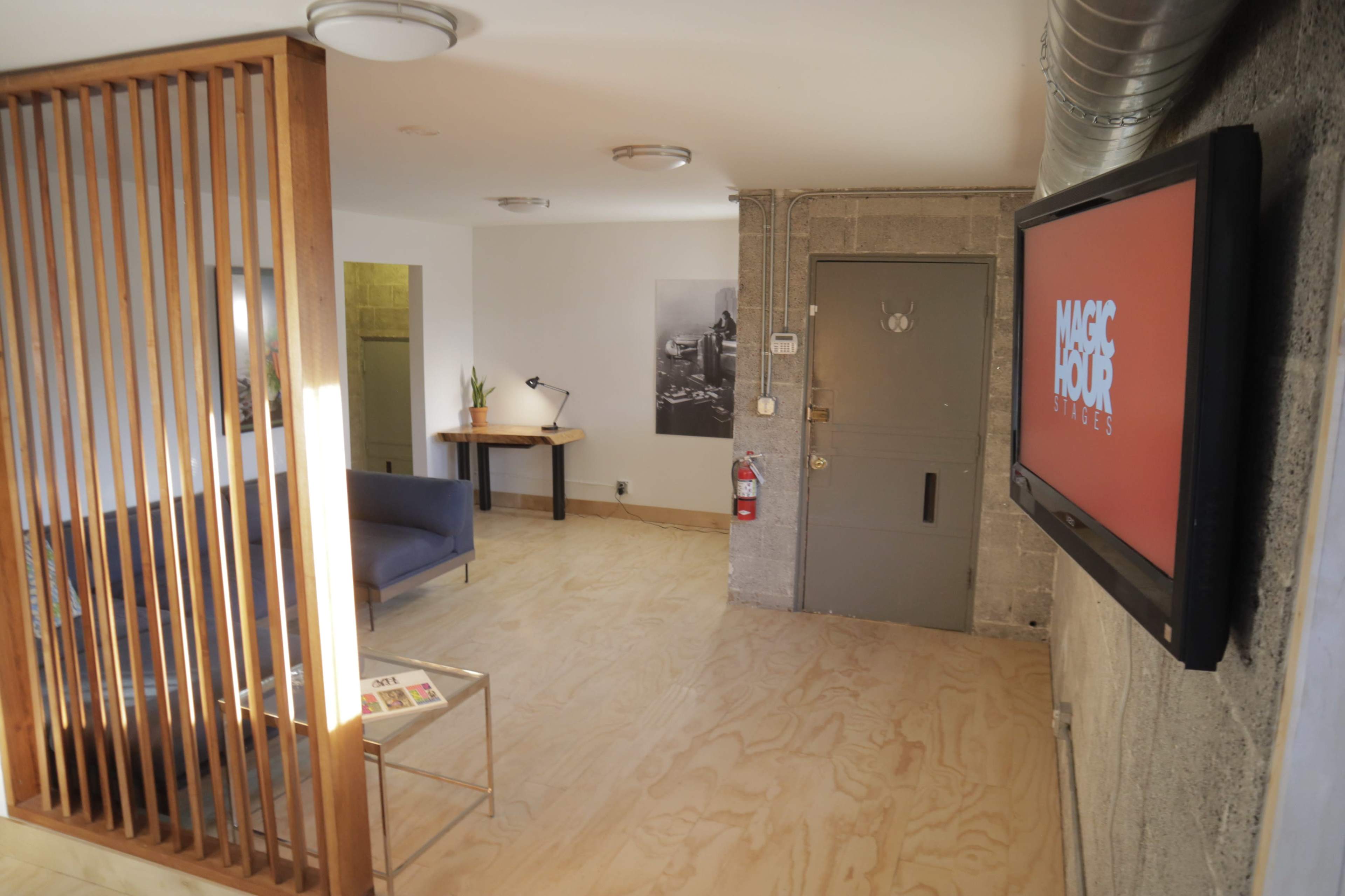 The image shows a modern interior space with a partitioned sitting area featuring a blue sofa, a coffee table with magazines, and a wall-mounted screen displaying a logo, alongside a concrete wall and an entry door.