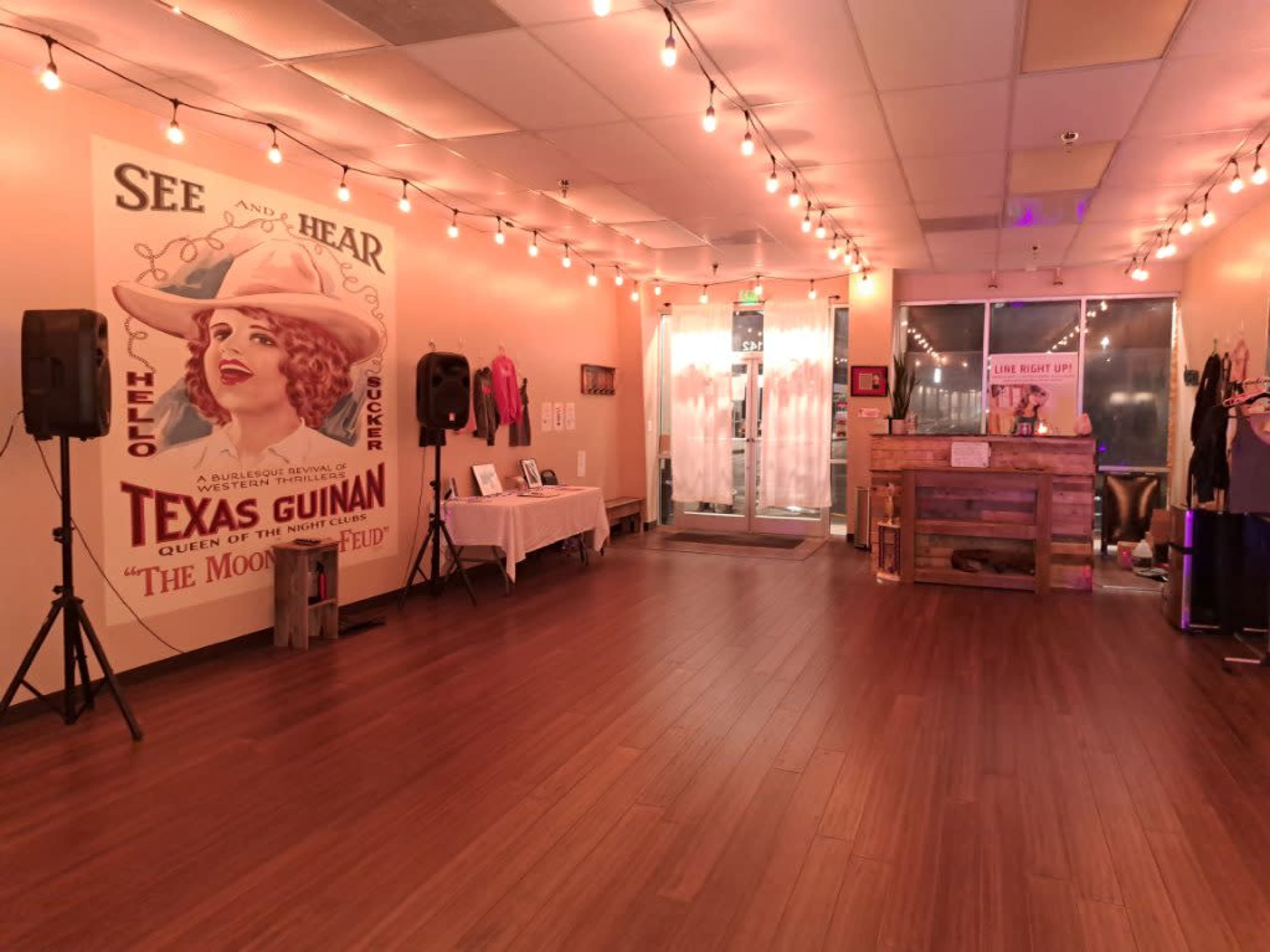 The interior of a well-lit venue features a vintage poster of Texas Guinan on the wall, wooden flooring, and a reception area with a rustic wooden counter.