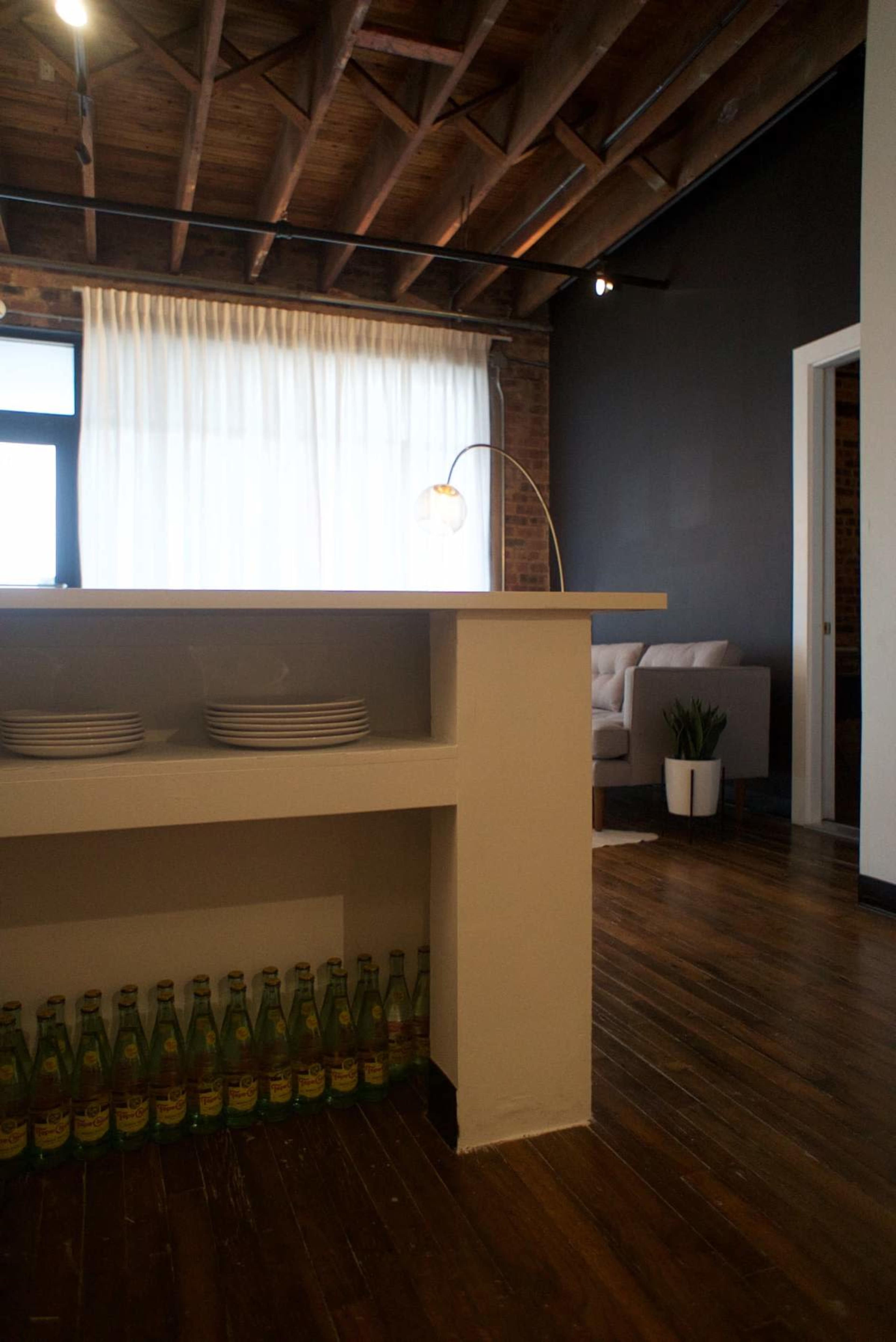 The image shows a modern interior space with a counter featuring stacks of plates and bottles lined up below, wooden flooring, and a seating area in the background.
