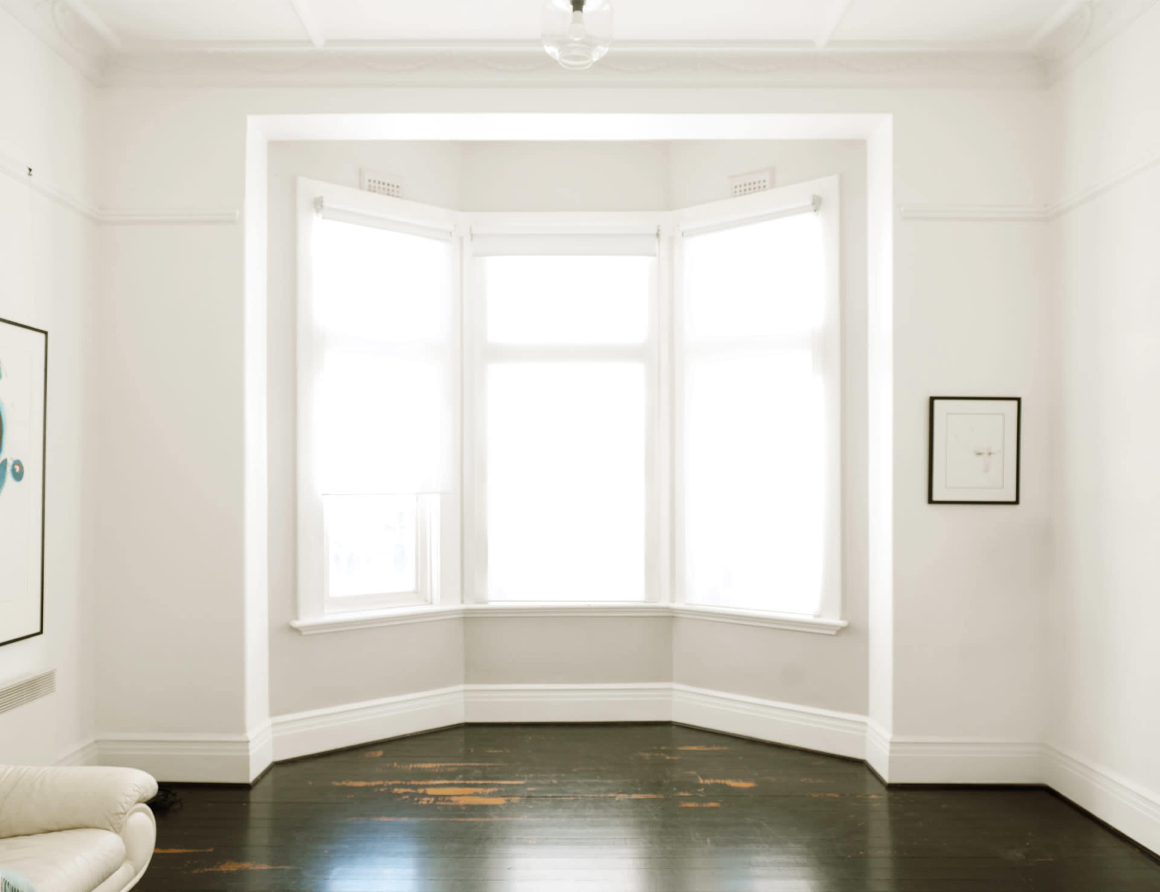 A bright, empty room features large windows with white curtains and dark wooden floors.