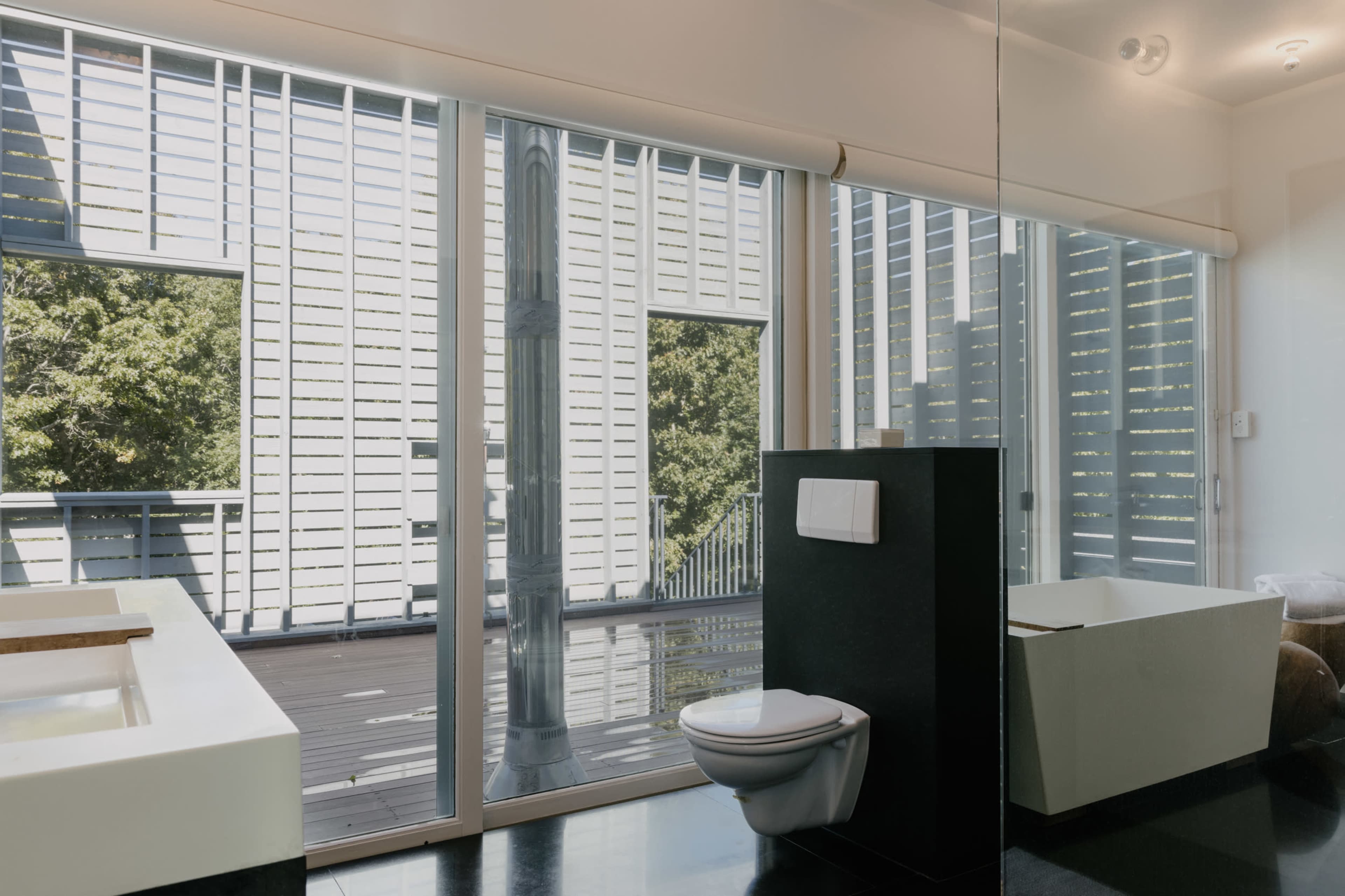 A modern bathroom with a freestanding bathtub, a toilet, and large glass windows that open to a deck overlooking greenery.