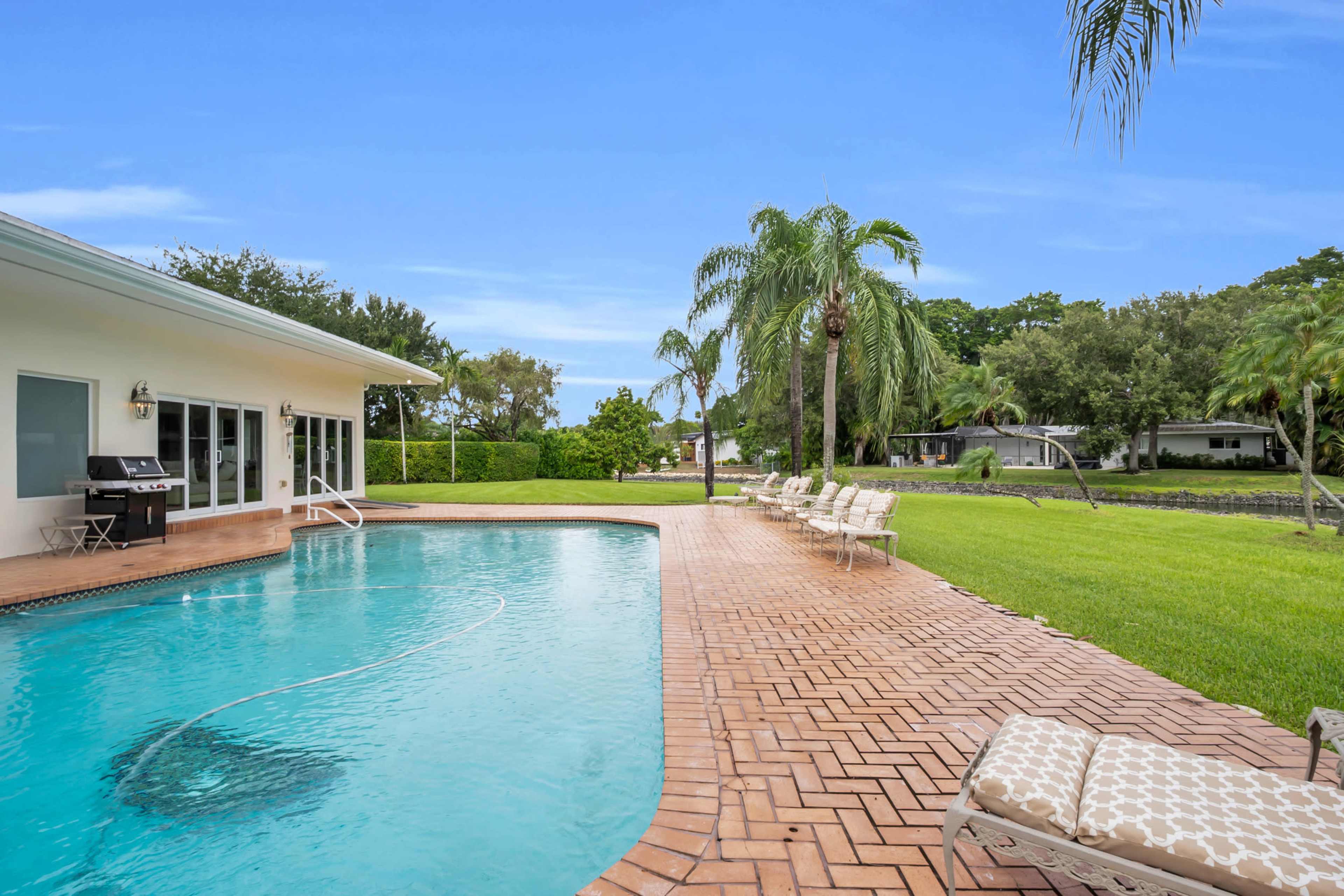 The image shows a swimming pool surrounded by a patio and lounge chairs, with palm trees and grassy areas in the background.