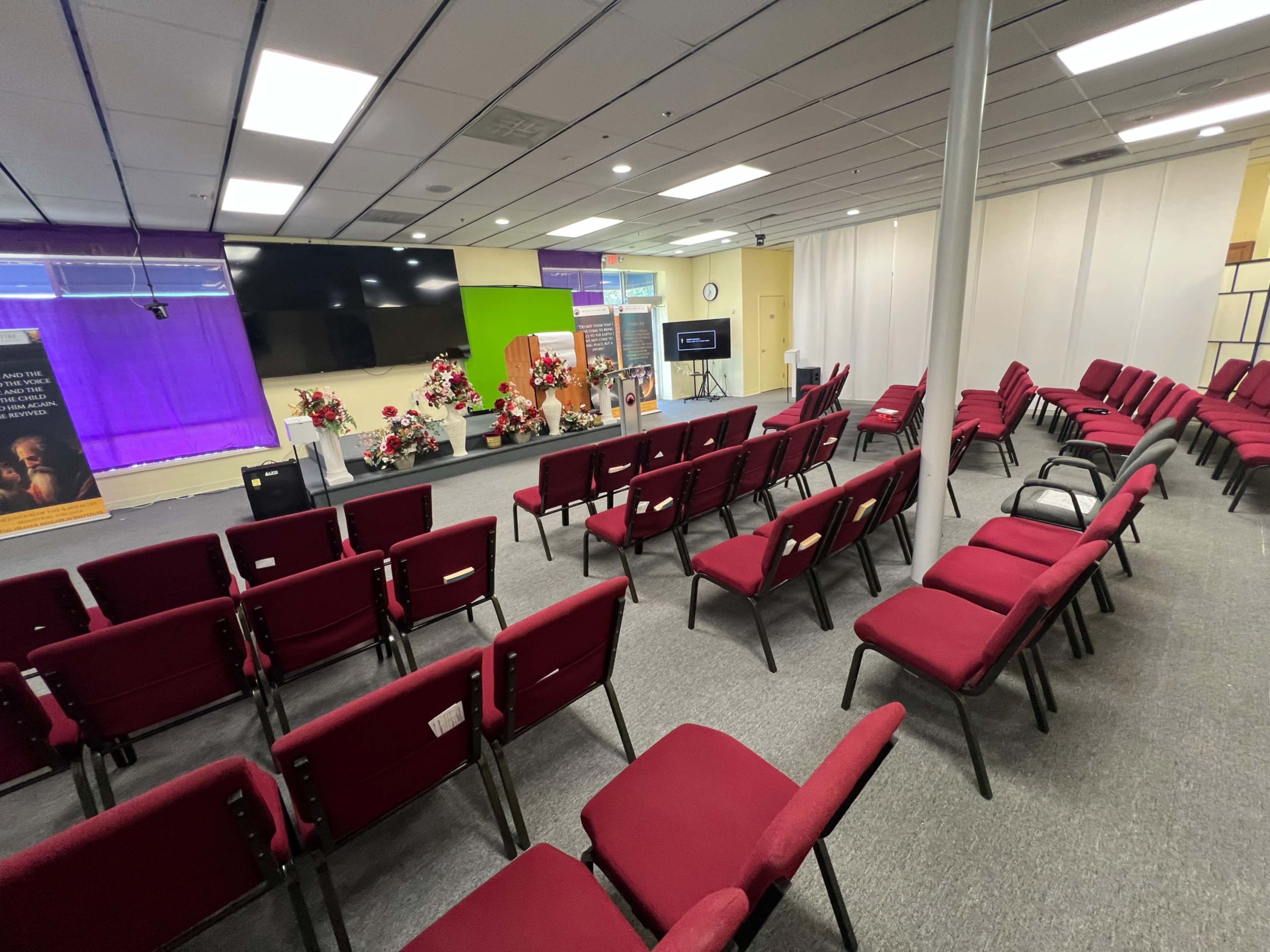 A spacious room with maroon chairs arranged in rows facing a stage with floral decorations and a large screen.