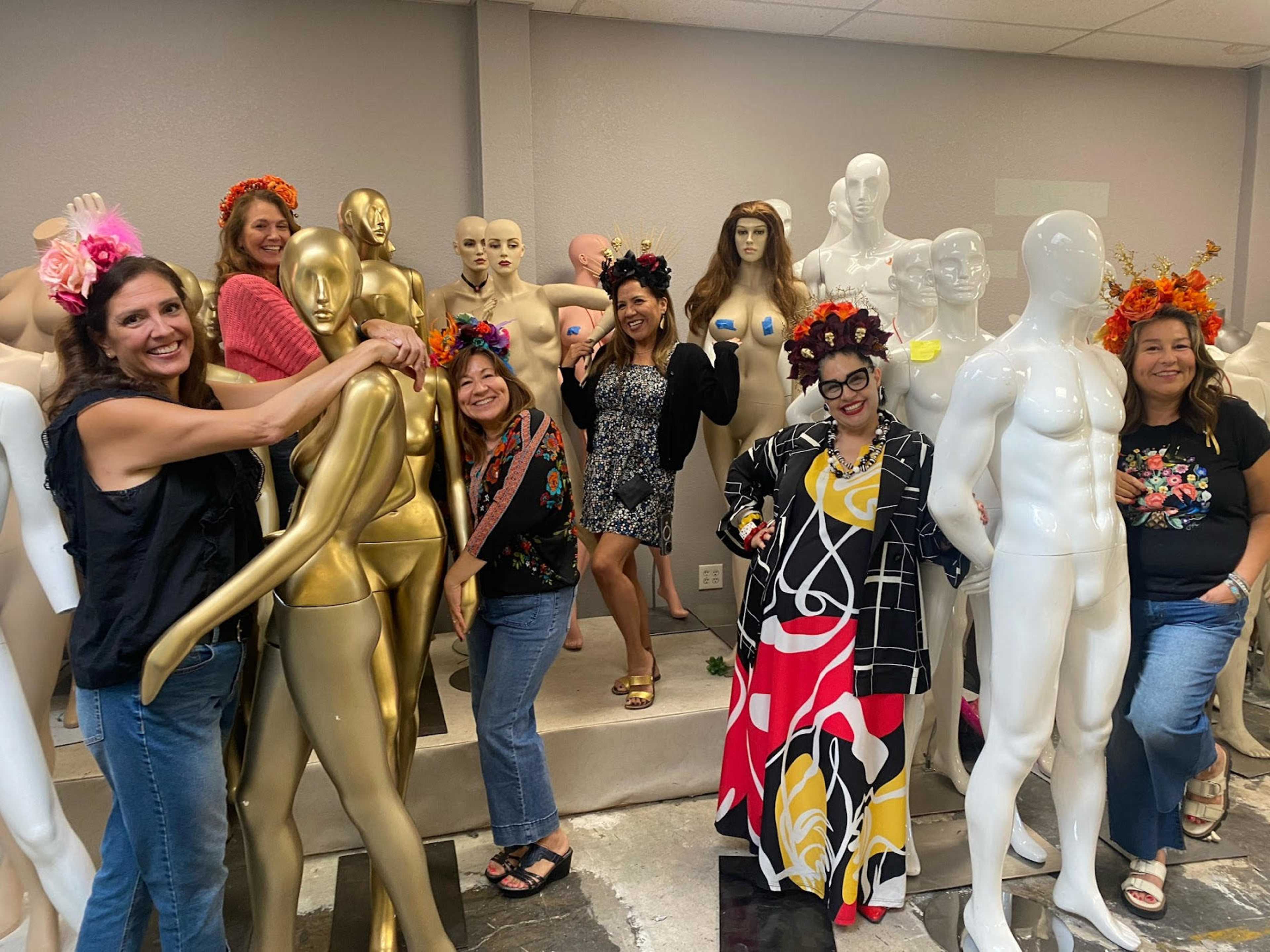 A group of seven women poses playfully amidst a display of mannequins, some adorned with floral headpieces.