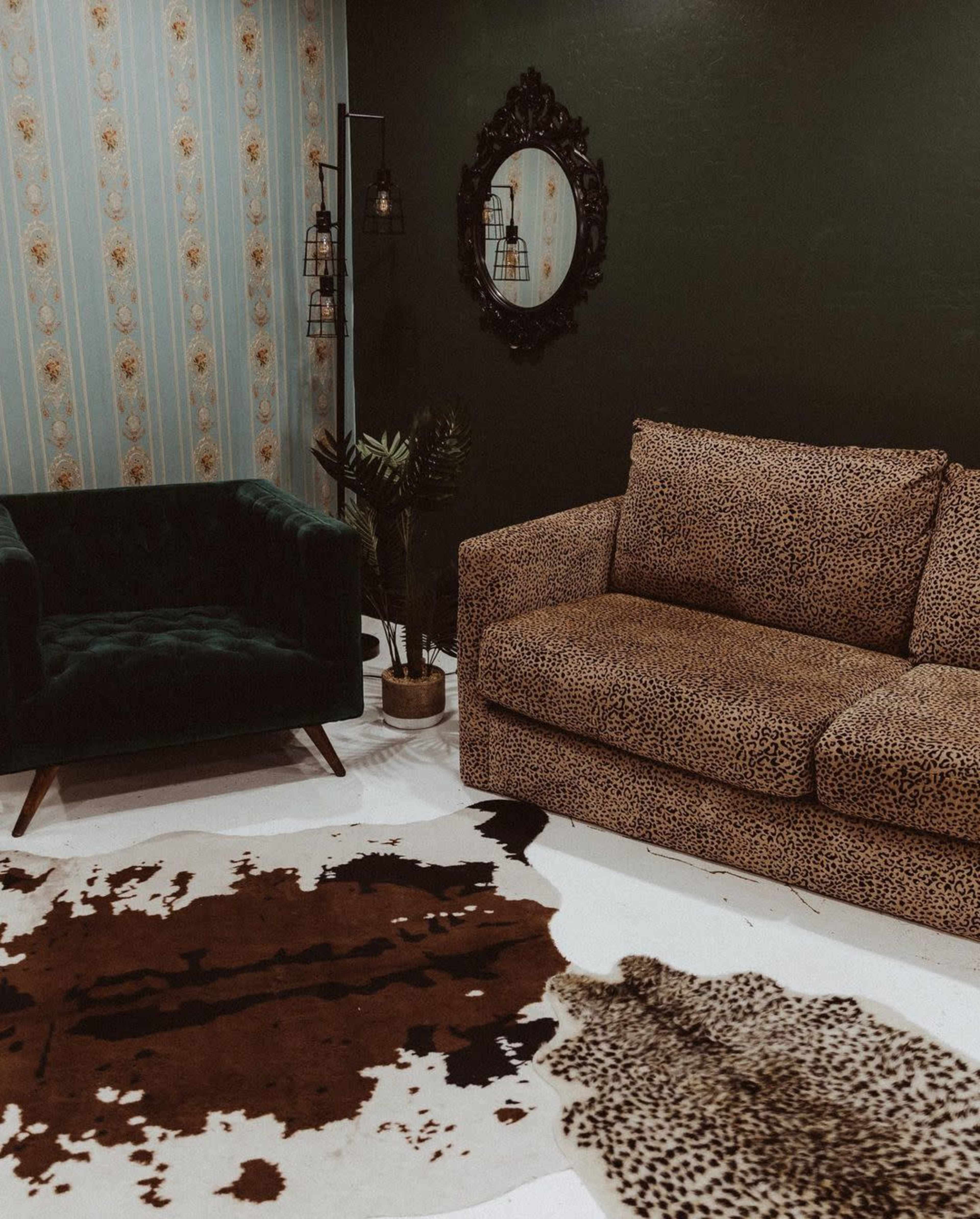 A living room with a leopard print sofa, a dark green armchair, and a cowhide rug on a light floor, surrounded by green walls and patterned wallpaper.