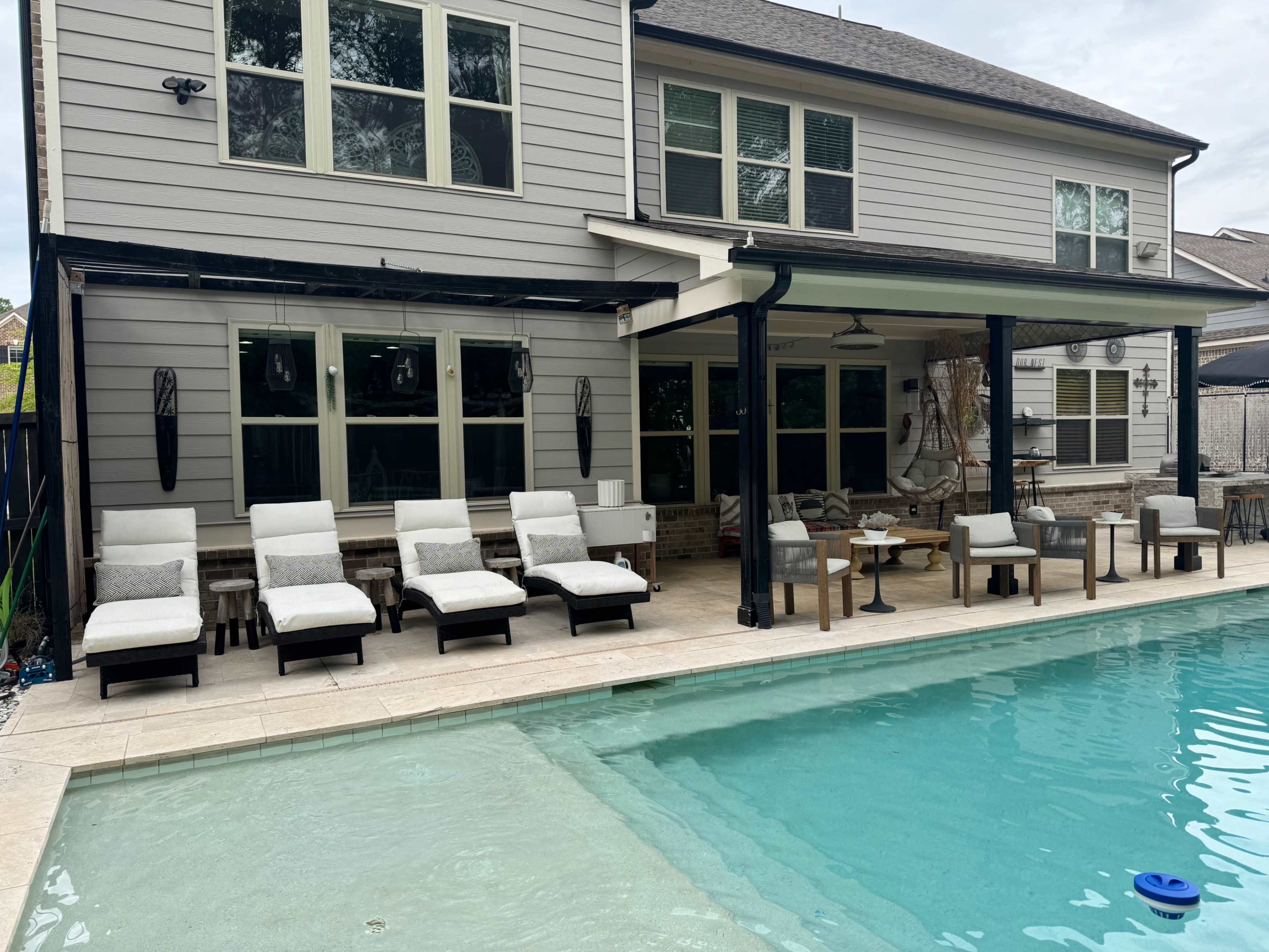 The image shows a residential backyard with a swimming pool, surrounded by lounge chairs and a covered patio area with tables and seating.